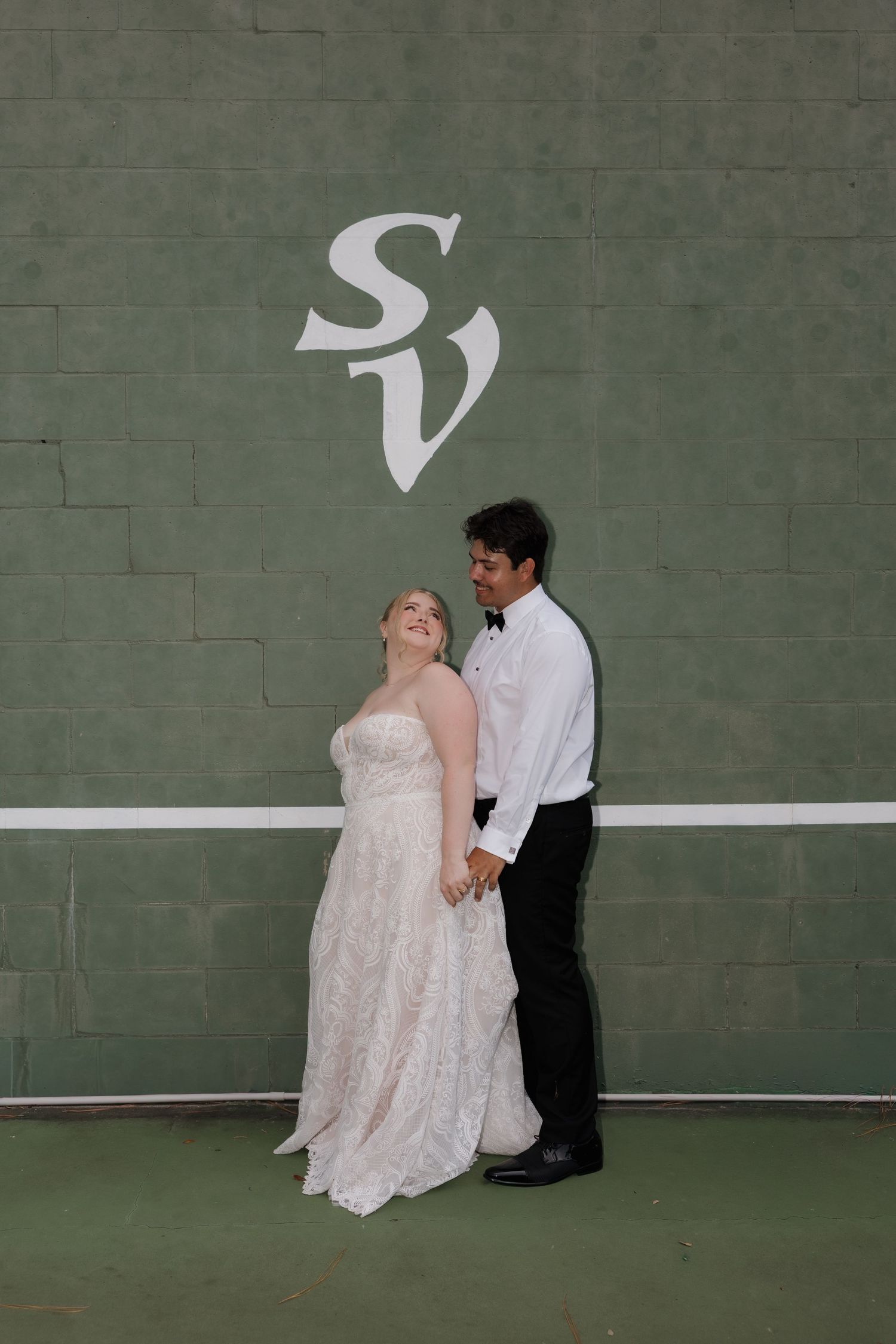 Bride in strapless lace gown and groom in tuxedo pose together in front of green wall with SV monogram.