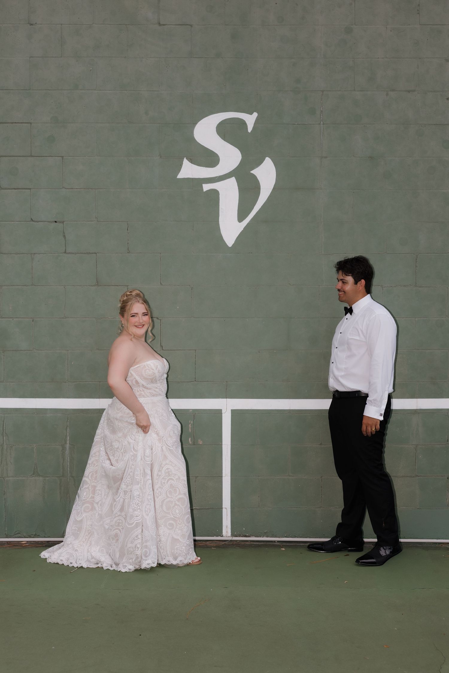 Bride and groom posing on tennis court in front of green wall with SV logo.