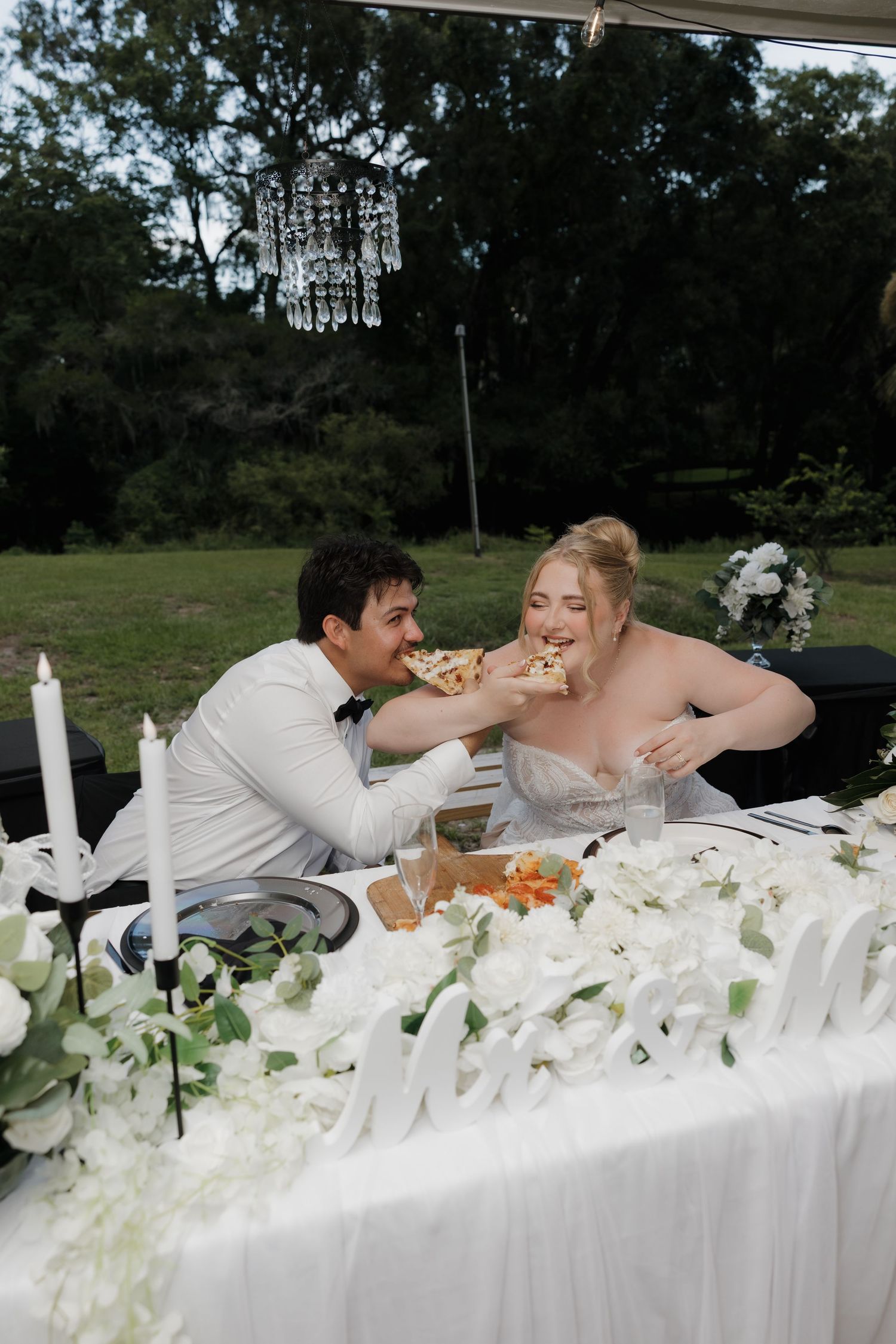 Bride and groom laugh while sharing pizza at their elegant outdoor wedding reception sweetheart table.
