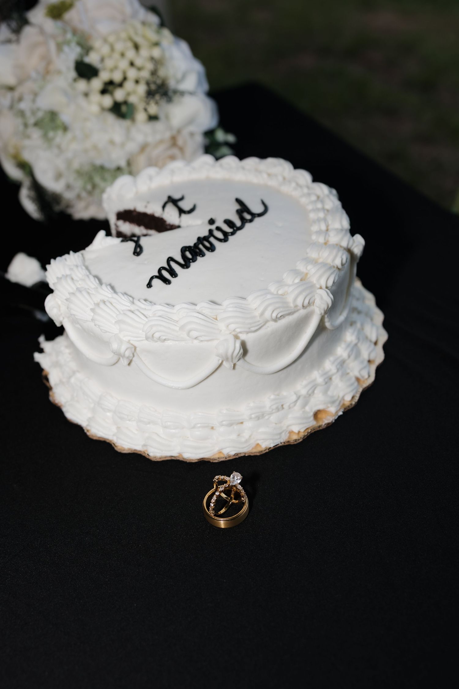 White two-tier wedding cake inscribed just married on black table with gold rings beside it.