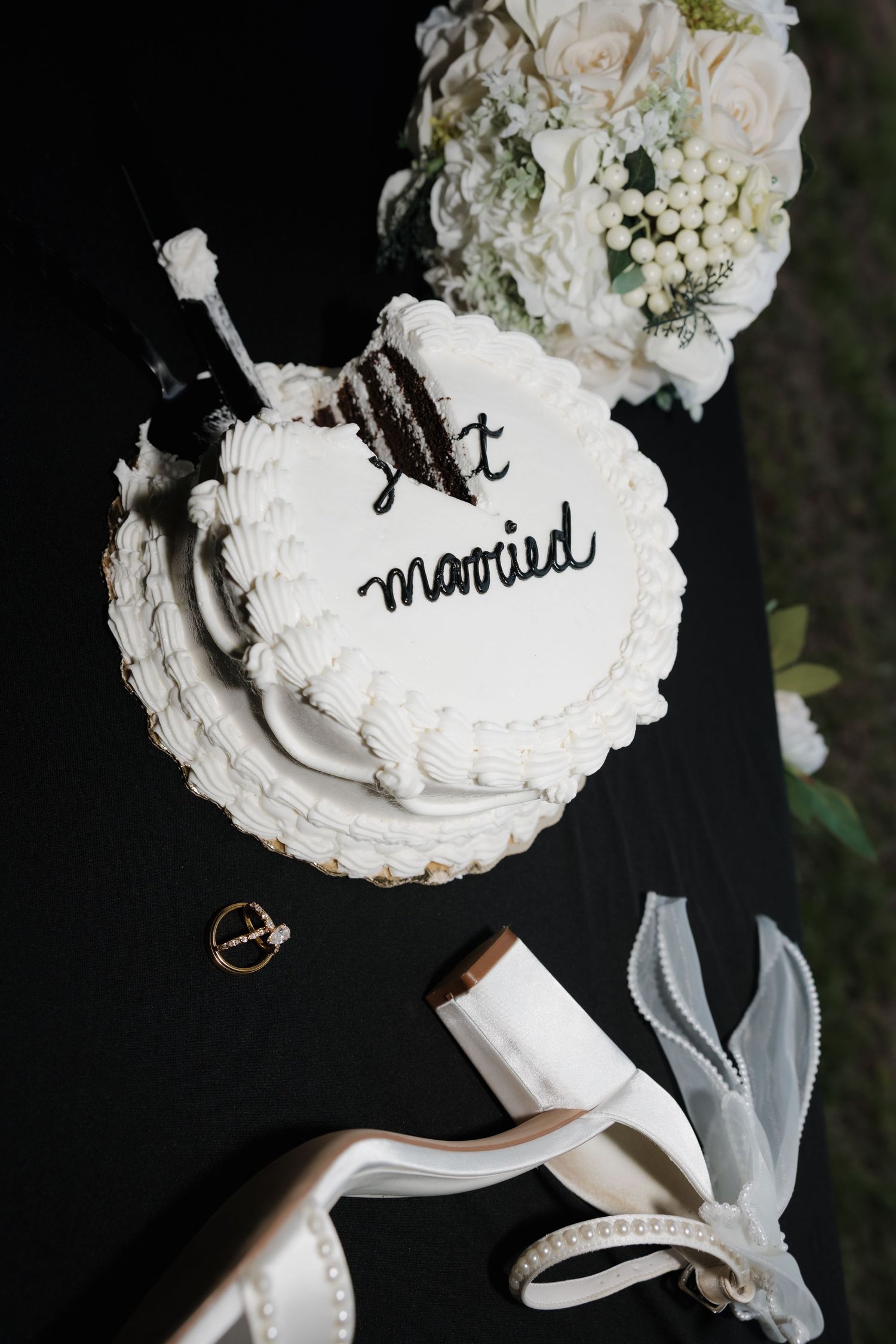 White wedding cake inscribed with just married beside bridal bouquet, gold rings, and heels on black table.