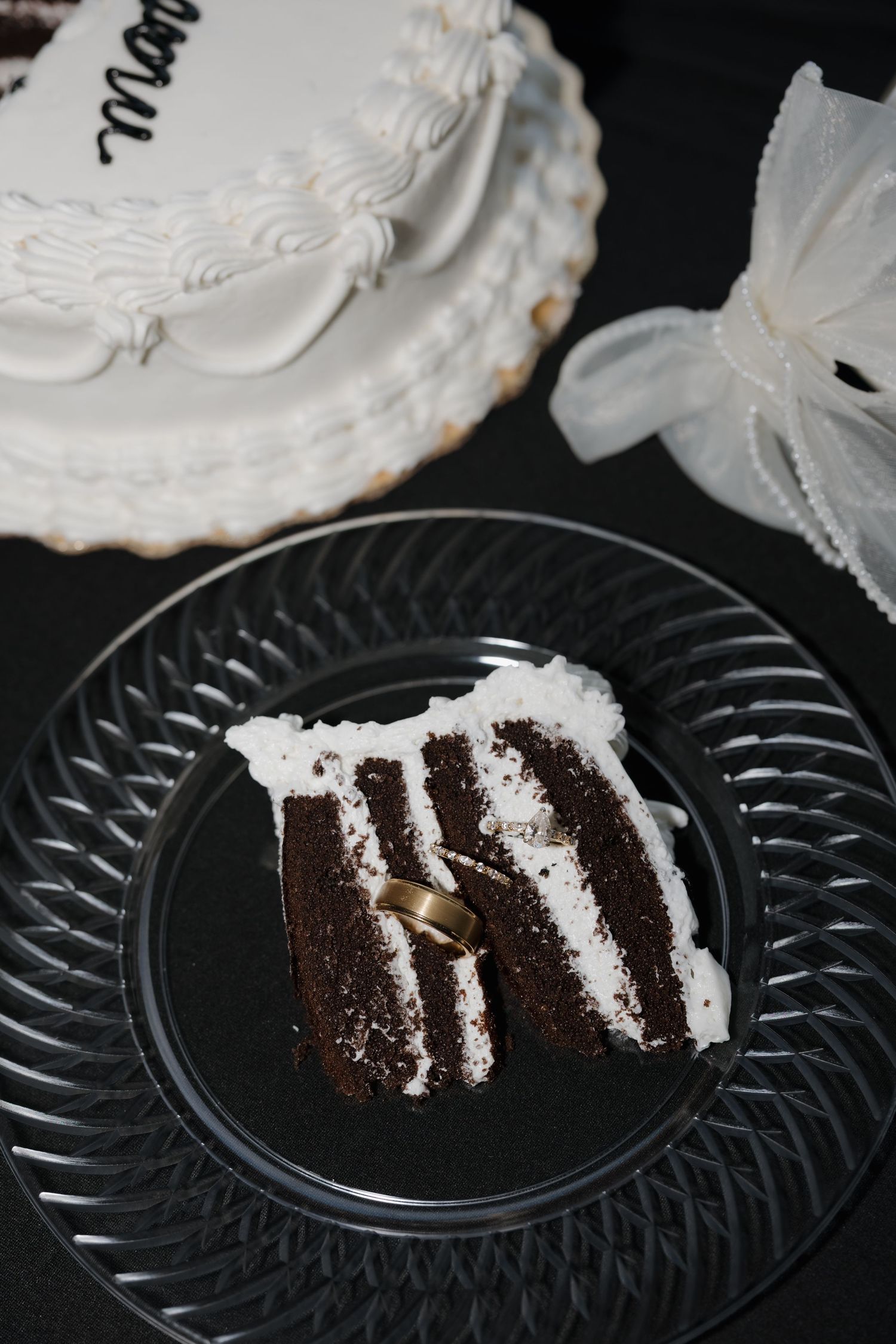 Slice of layered chocolate cake with white frosting on glass plate beside tiered white wedding cake on black background.