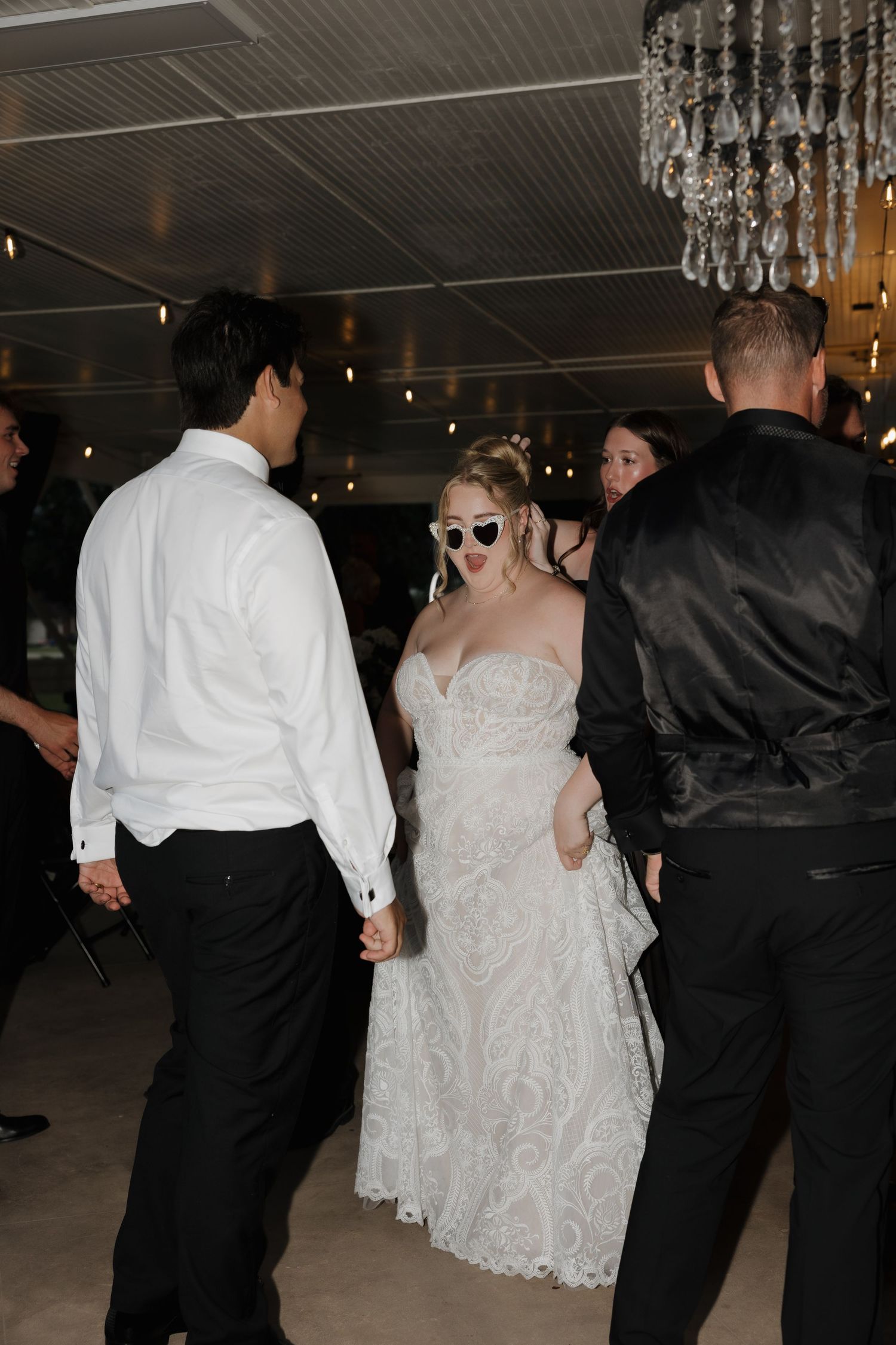 Bride in lace gown wearing heart-shaped sunglasses dancing with guests at wedding reception.