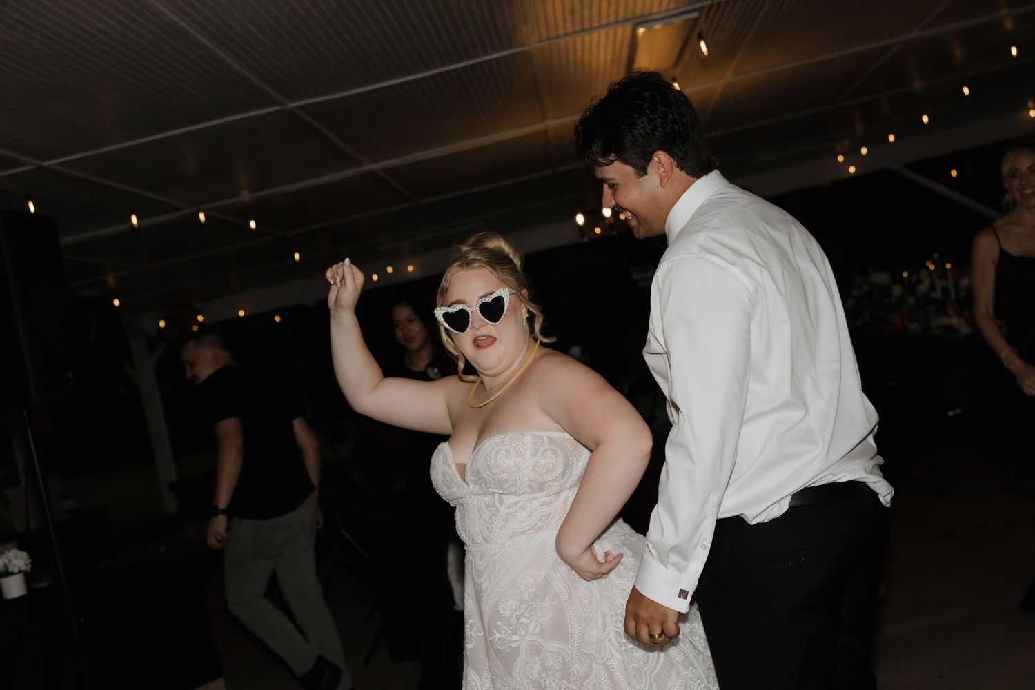 Bride in lace gown and white sunglasses dancing with groom in white shirt at outdoor wedding reception.