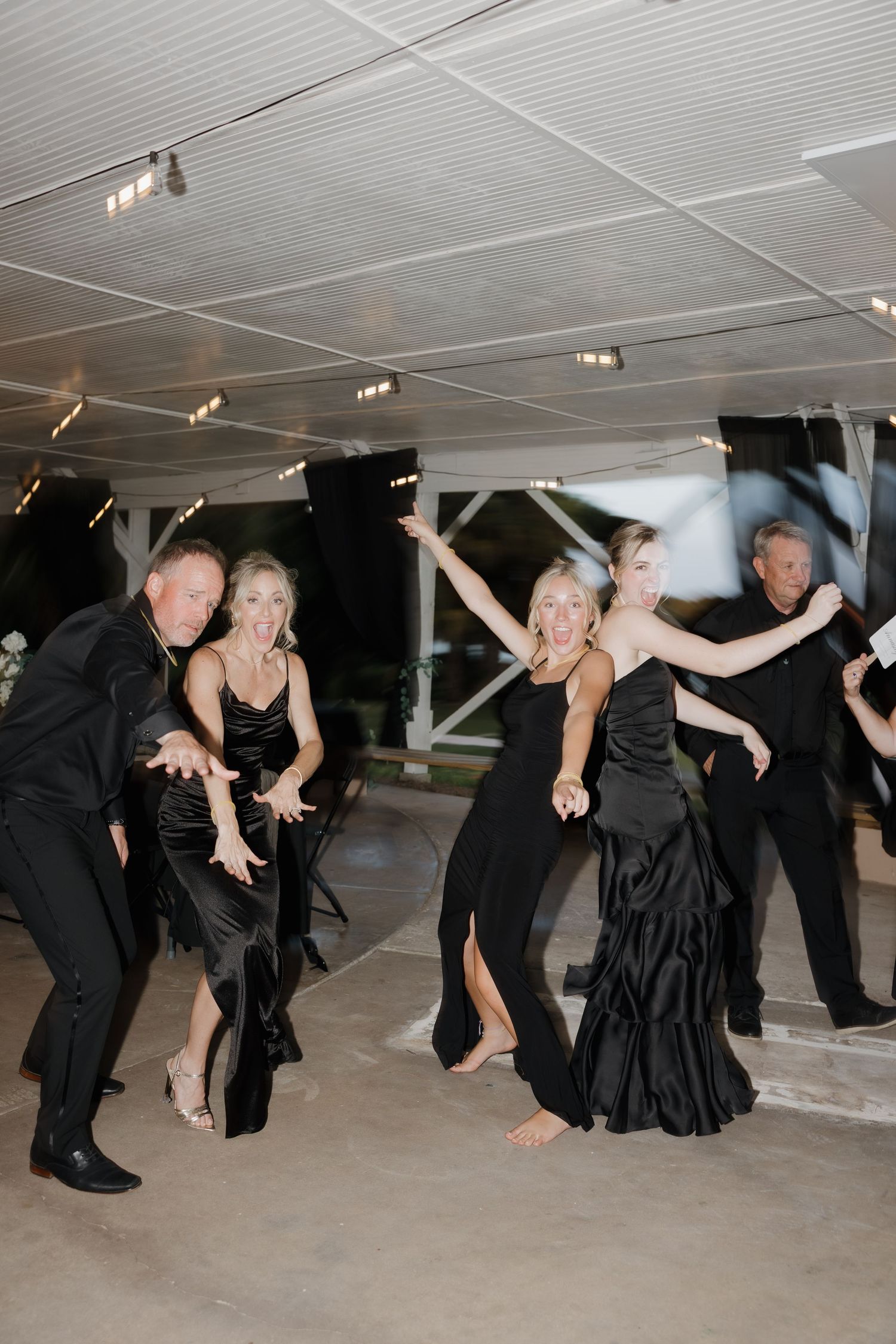 Wedding guests in black formal attire dancing energetically under covered outdoor reception venue.