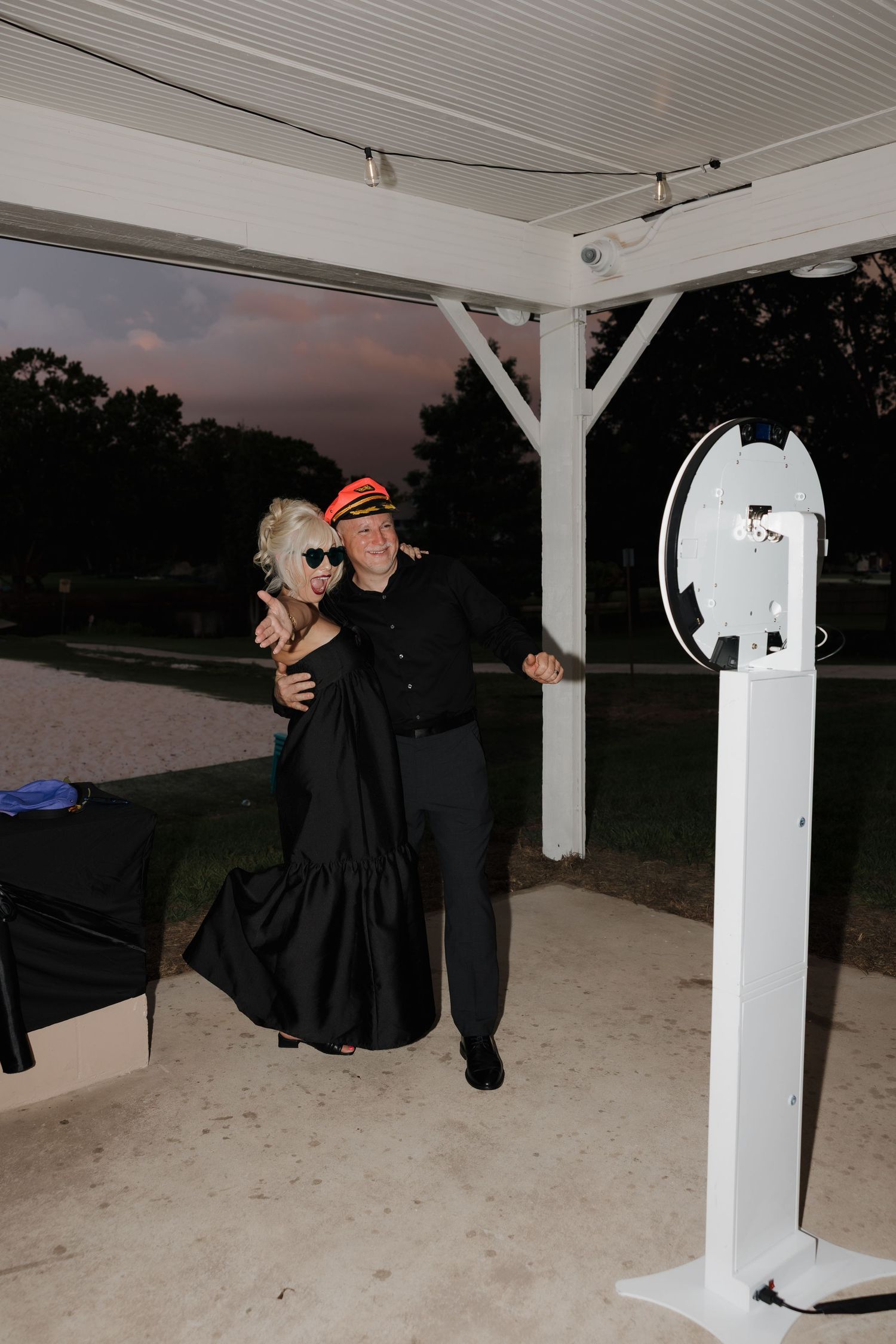 Couple in black formal attire posing together at an outdoor photo booth under a gazebo at dusk.