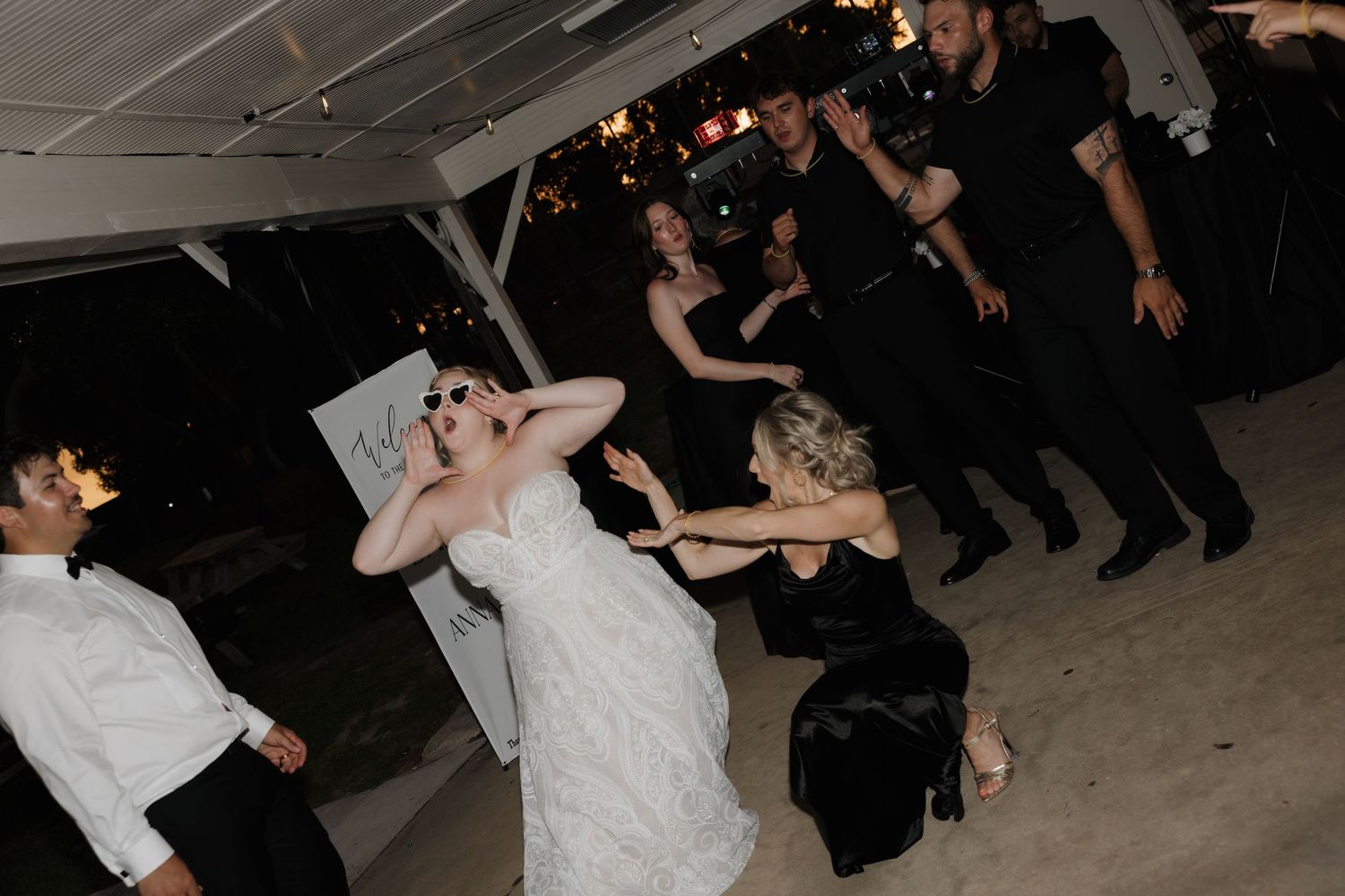 Bride in white gown dancing joyfully with wedding guests at an outdoor evening reception celebration.