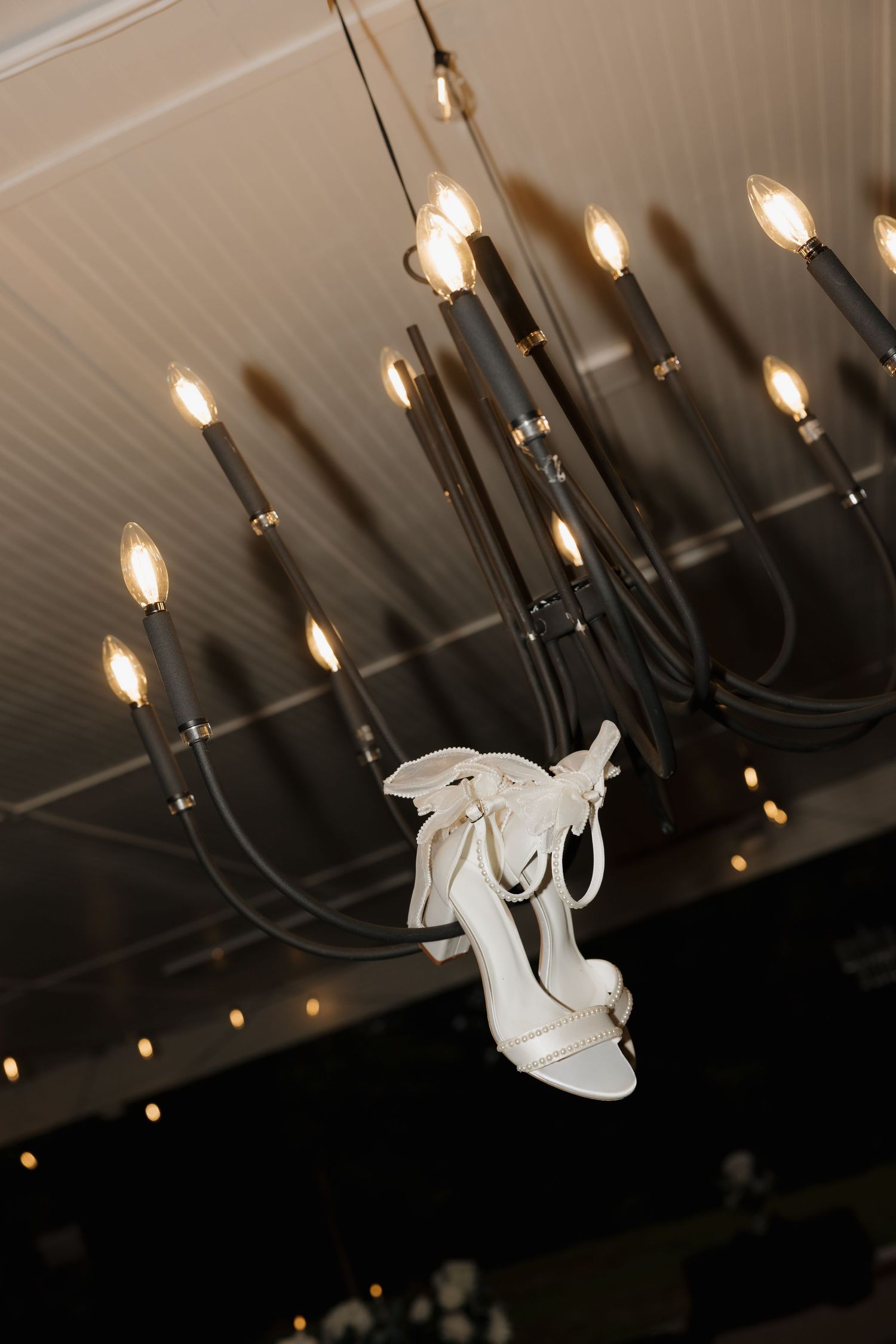 White bridal high heel shoe hanging from an elegant black candelabra chandelier with lit bulbs.