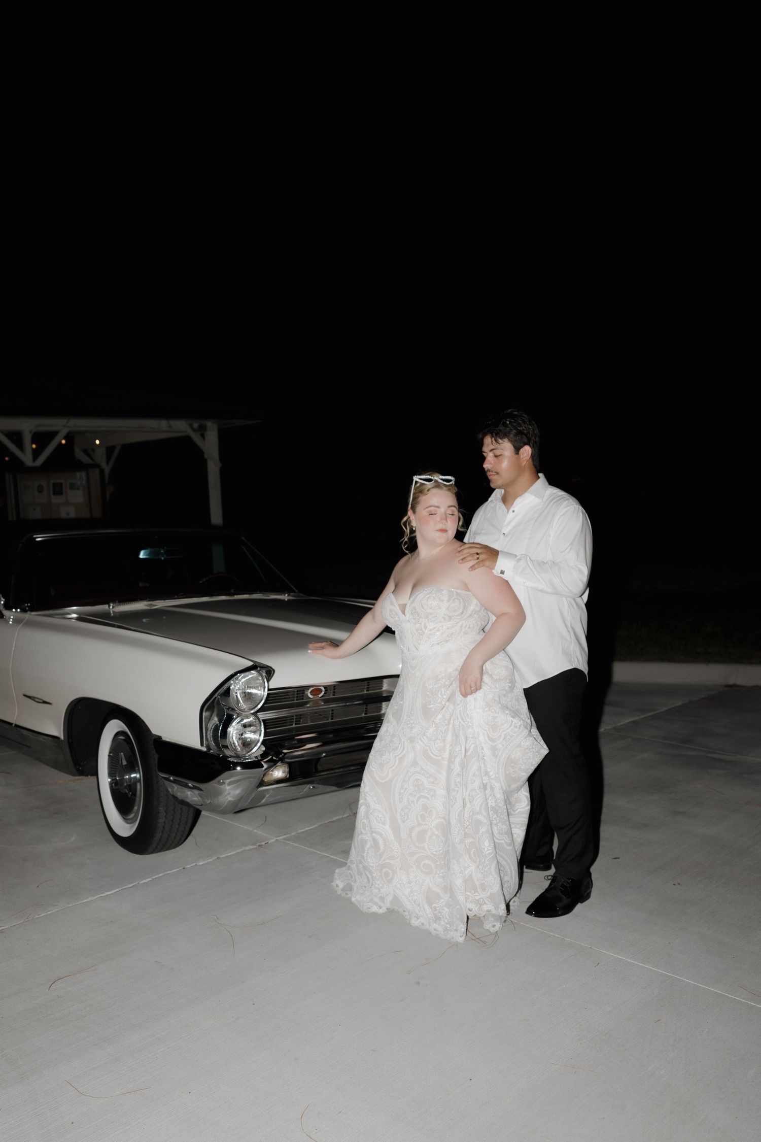 Bride in white lace gown and groom in white shirt pose beside a classic white convertible at night.