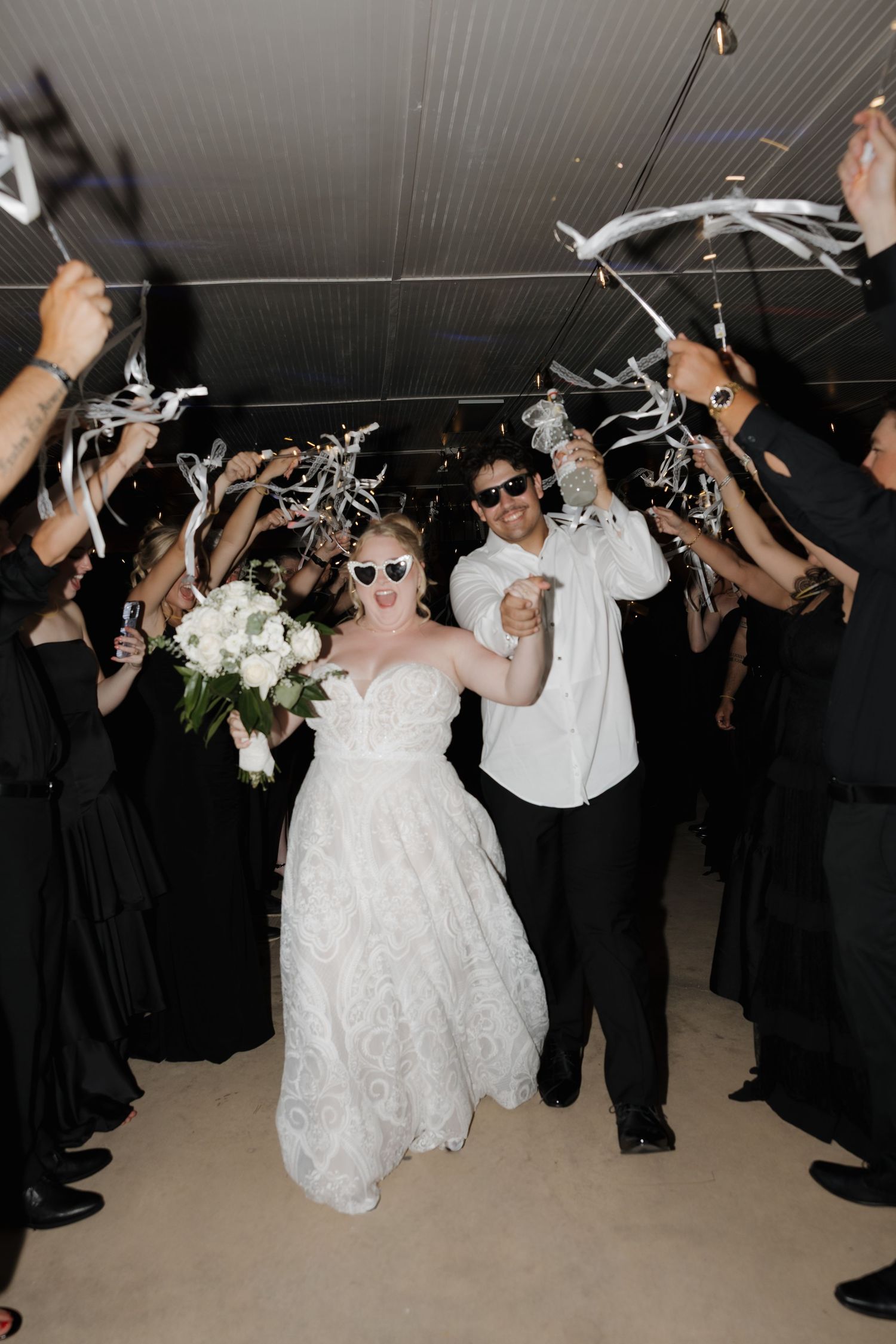 Bride and groom exit wedding reception in sunglasses through sparkler tunnel with guests in black attire.