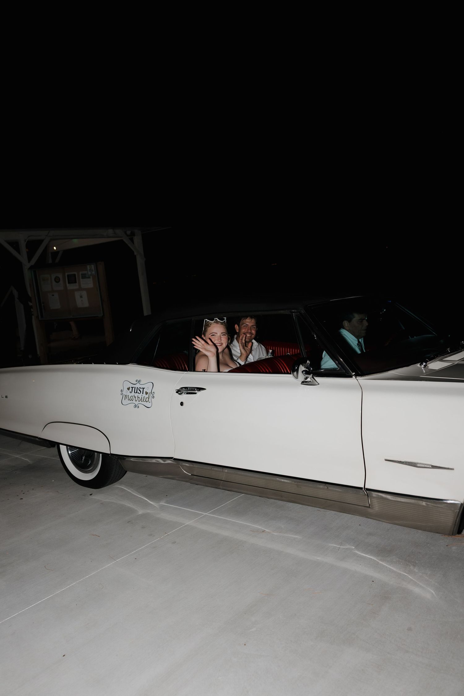 Couple smiling in a classic white vintage convertible car at night, with red interior seating.