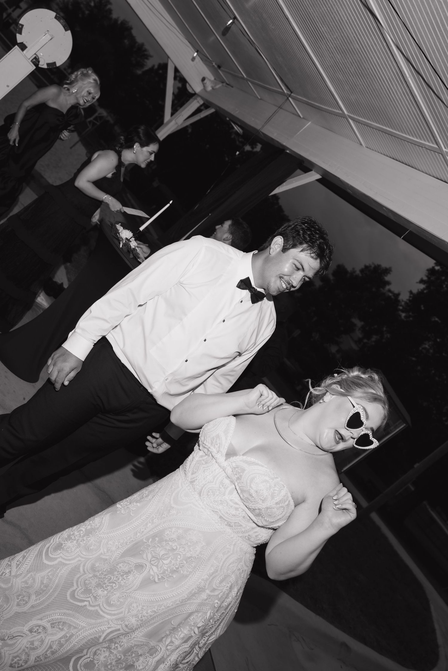 Bride in lace gown wearing sunglasses dances with groom in white shirt and bow tie at wedding reception.