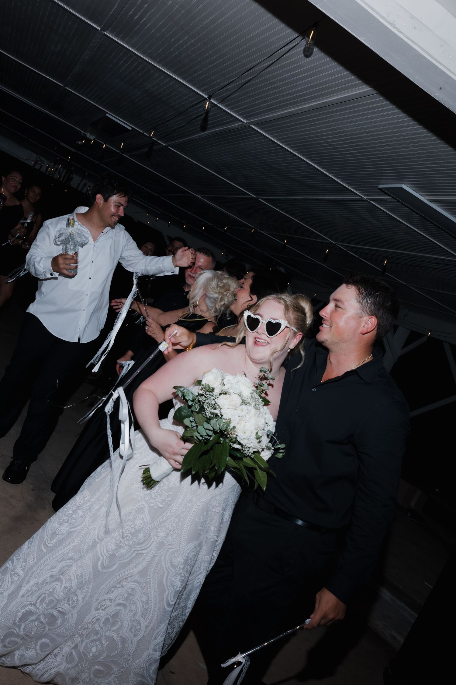 Bride in lace gown and heart sunglasses holds white bouquet while walking with groom at wedding reception.