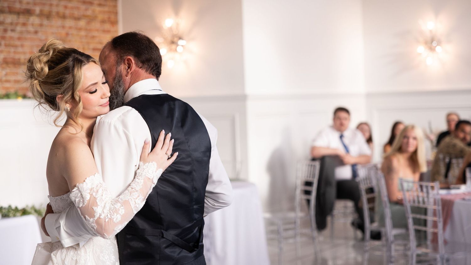 A bride and groom share their first dance at an elegant wedding reception with guests seated in the background.