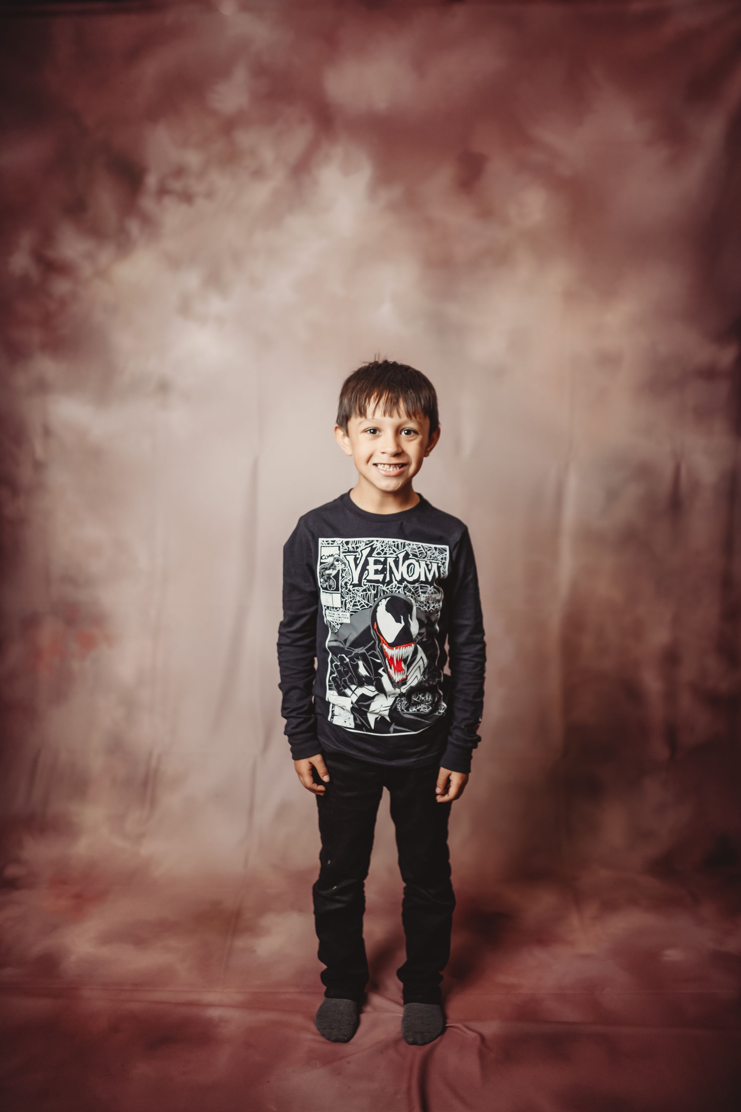 Young boy smiling in a Venom Marvel sweatshirt and black jeans, posing against a moody brown studio backdrop.