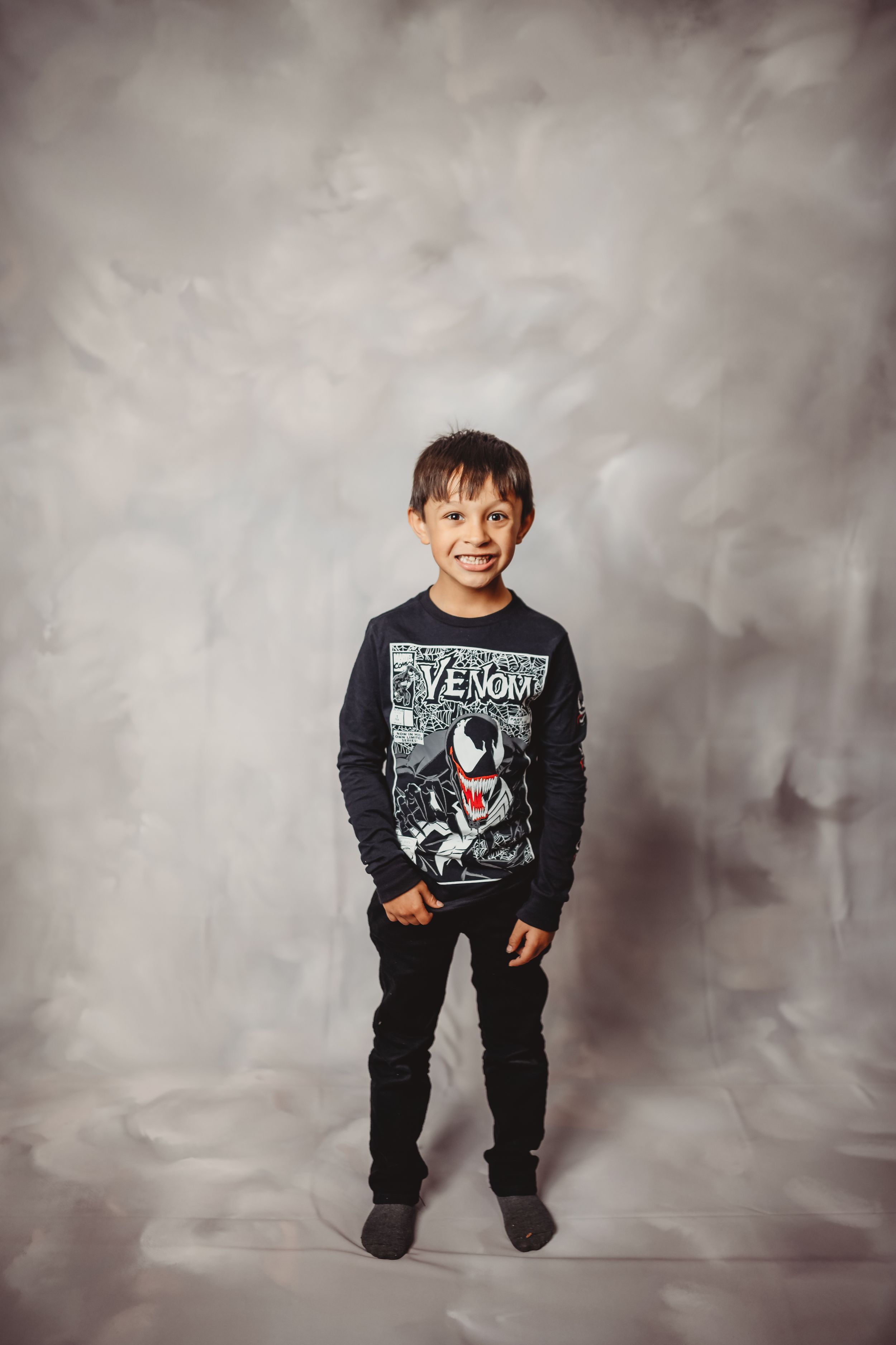 Young boy smiling in Venom Marvel shirt and black pants standing against gray studio backdrop.