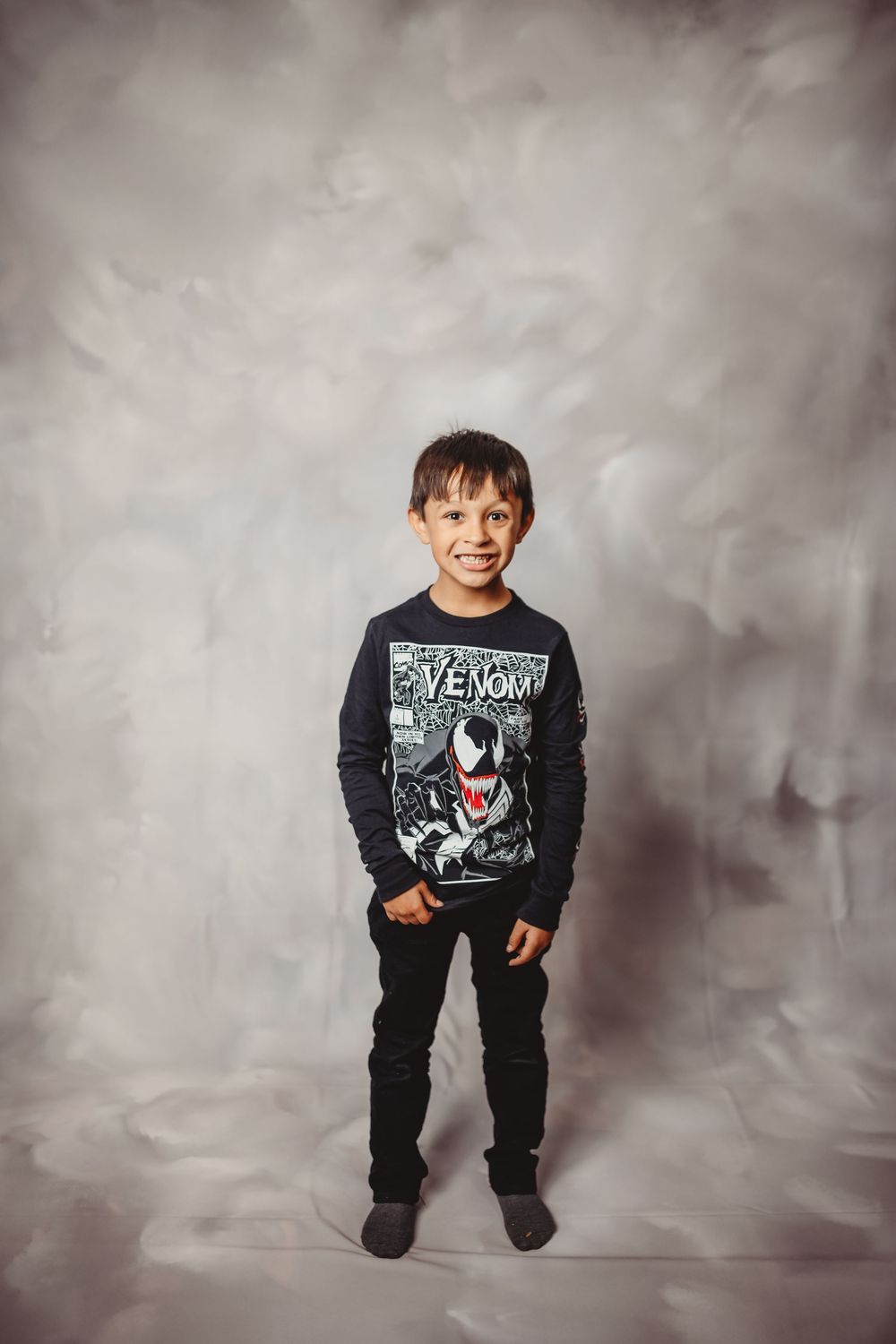 Young boy smiling in Venom Marvel shirt and black pants standing against gray studio backdrop.