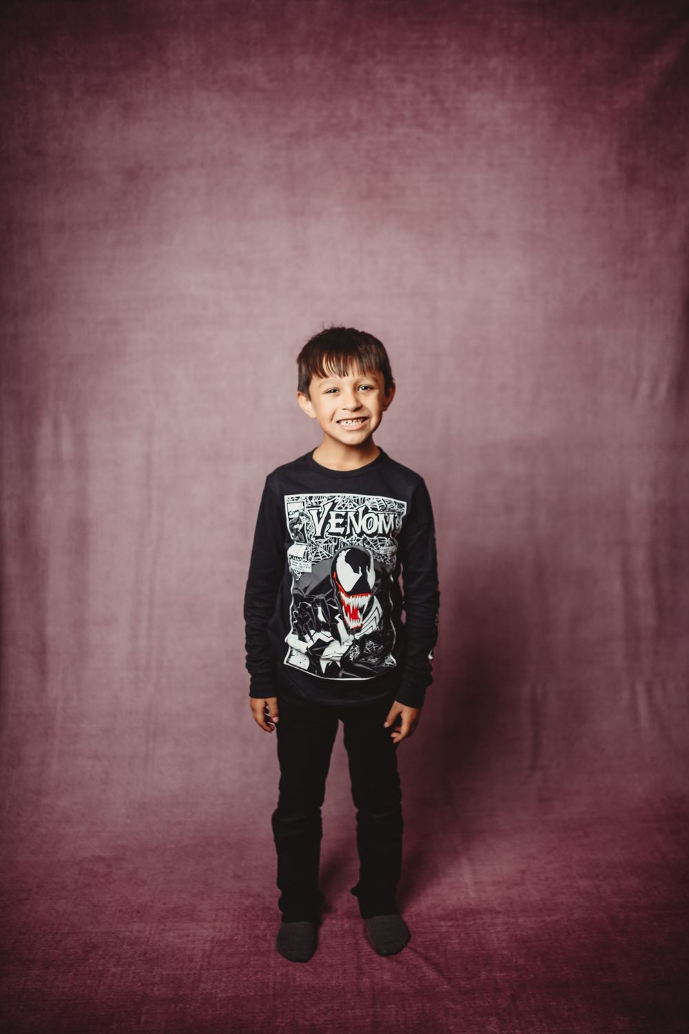 Young boy smiling in a Venom graphic long-sleeve shirt and black pants, standing against a purple backdrop.