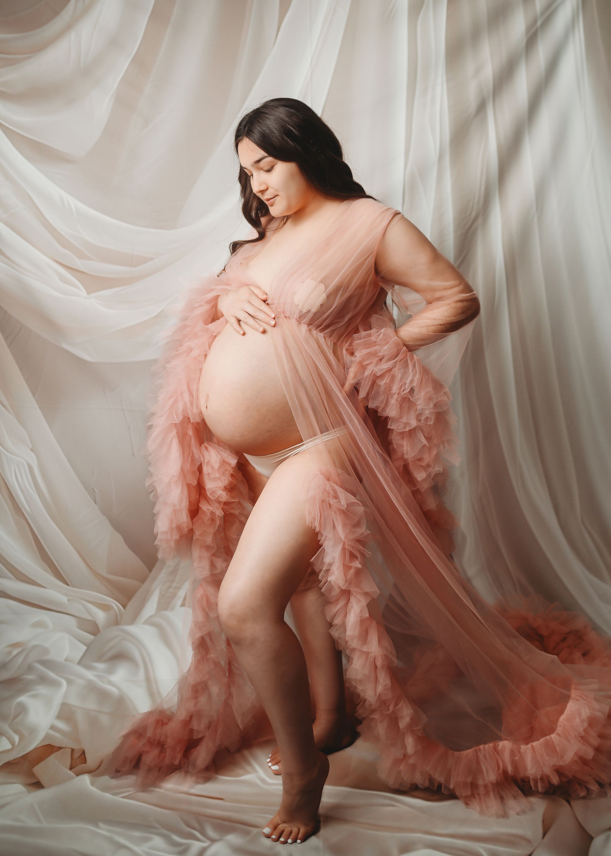 Pregnant woman in pink tulle maternity robe posing gracefully against soft white draped fabric backdrop.