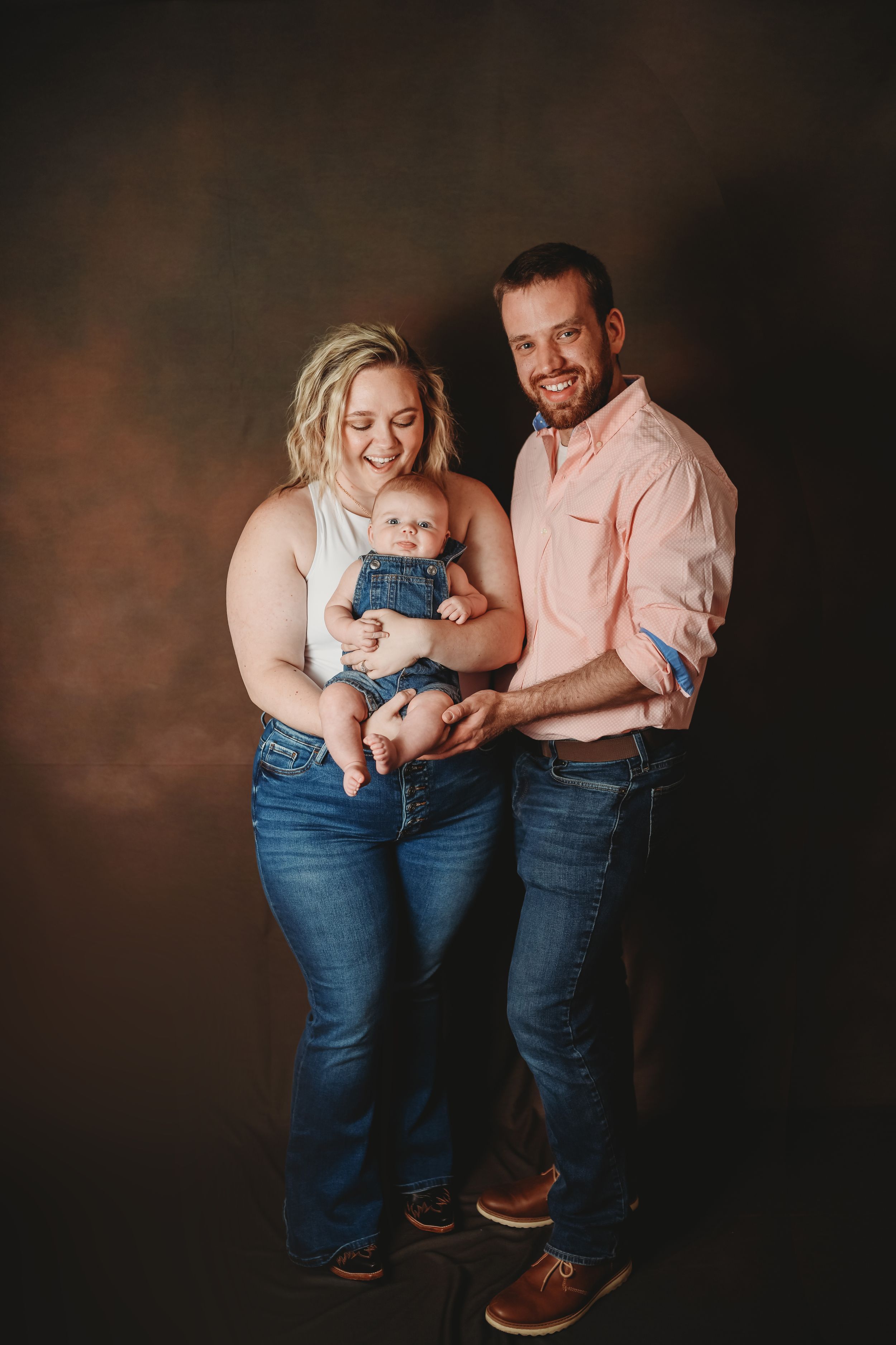 Happy family portrait with parents holding their baby in denim overalls against a brown studio backdrop.
