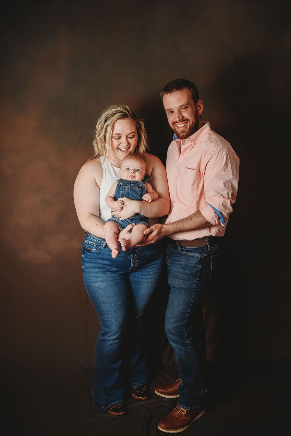 Happy family portrait with parents holding their baby in denim overalls against a brown studio backdrop.