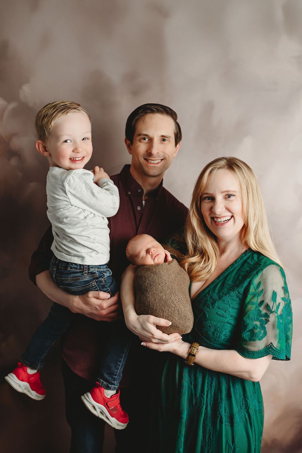 Happy family of four posing in studio, parents holding toddler boy and swaddled newborn baby, mom in green dress.