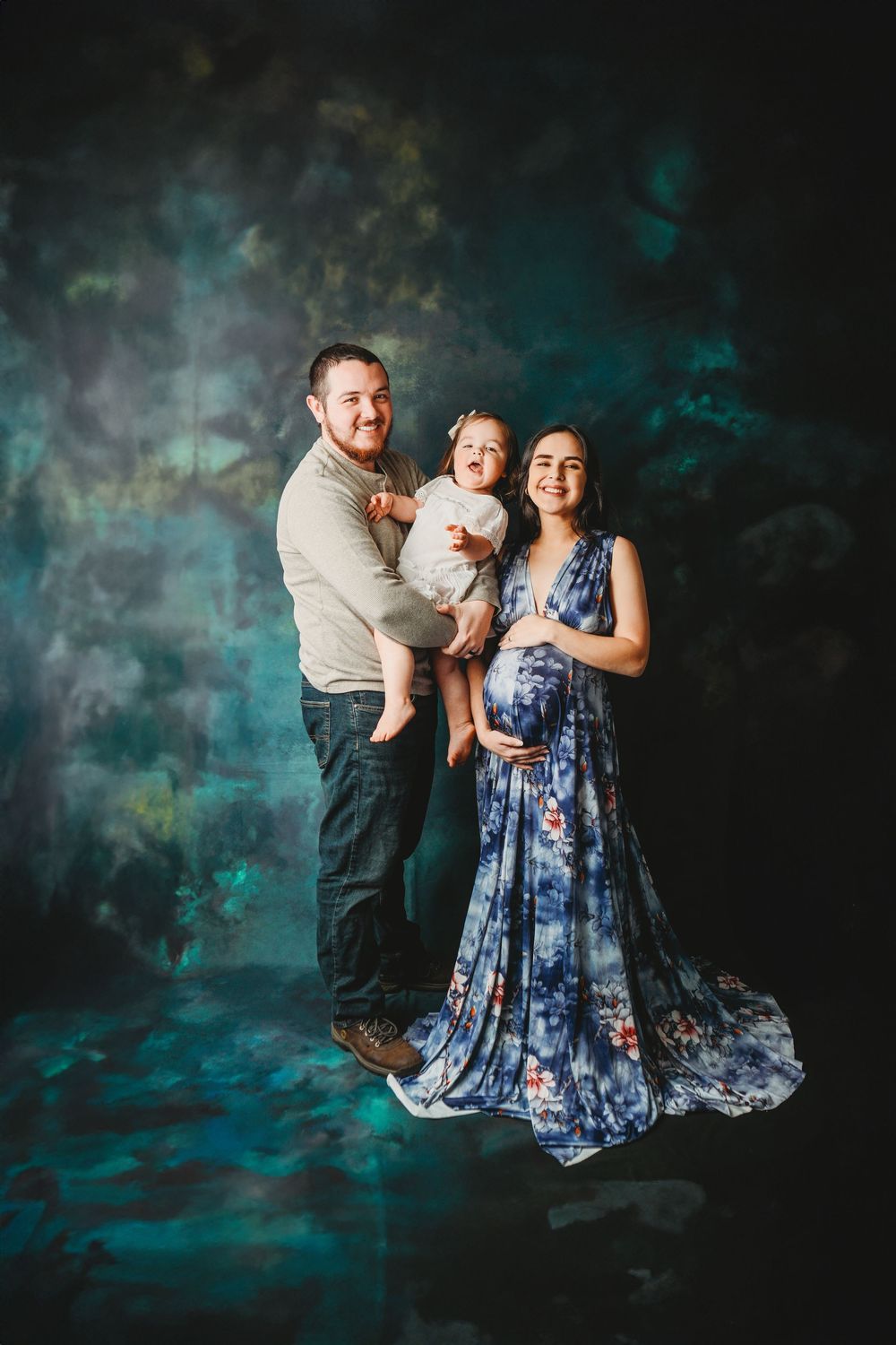 A happy family maternity photo with a man holding a baby girl and a pregnant woman in a blue floral dress.