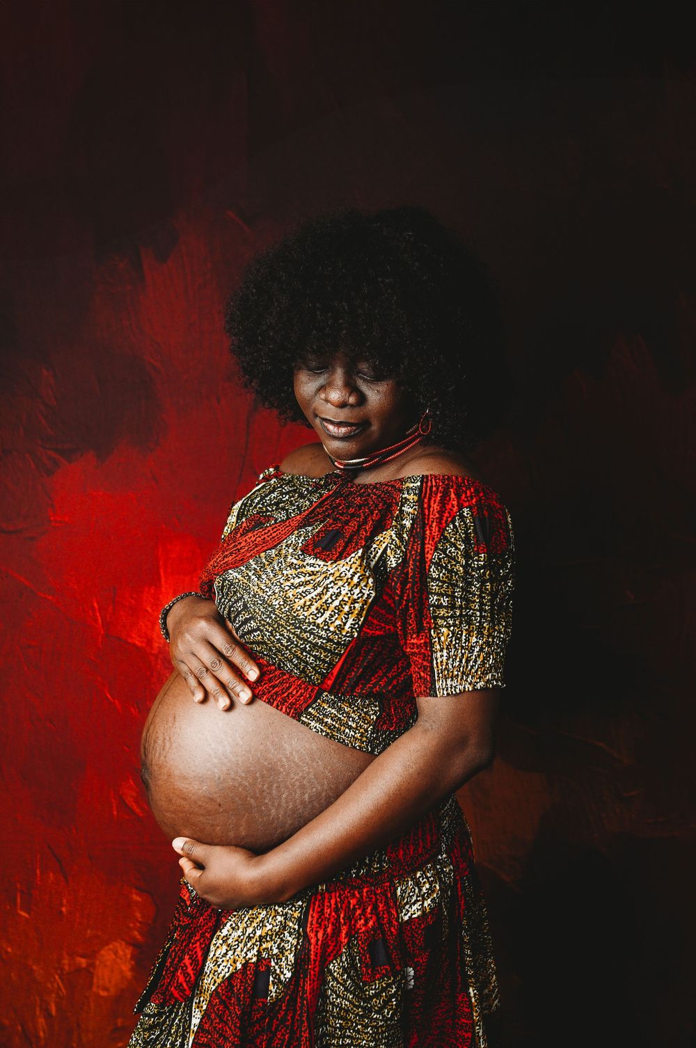 Pregnant Black woman in colorful African print dress cradling baby bump against dramatic red background.