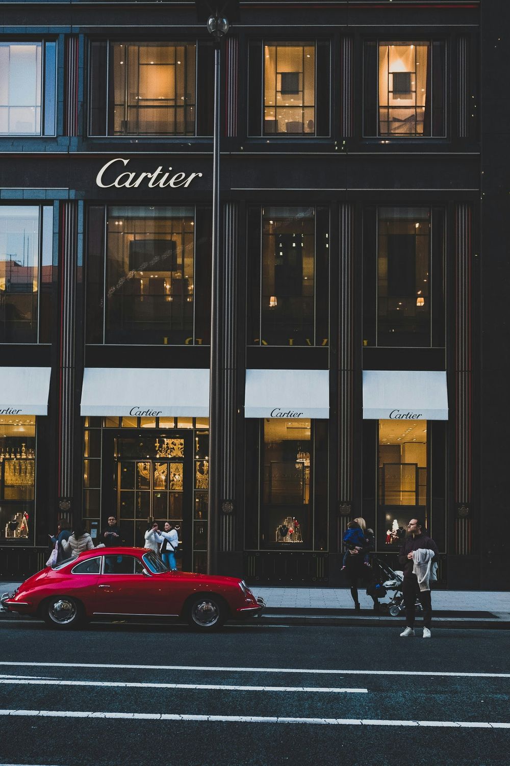 A vintage red Porsche parked outside a luxury Cartier store with pedestrians walking by on a city street.