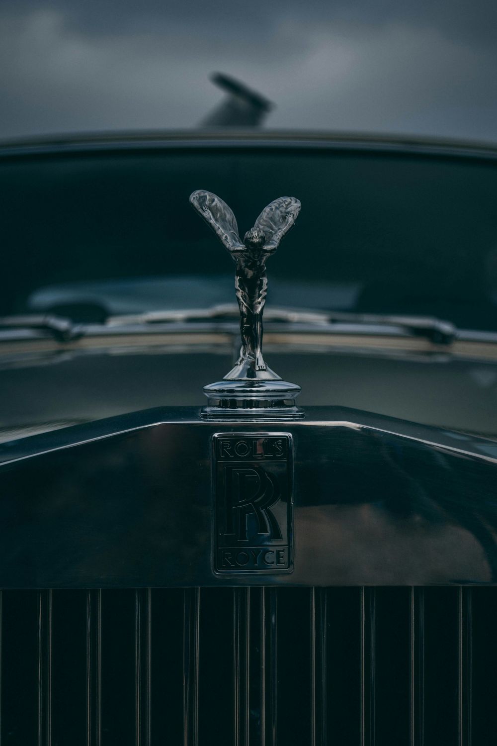 Close-up of a Rolls-Royce Spirit of Ecstasy hood ornament on a dark luxury car under overcast skies.