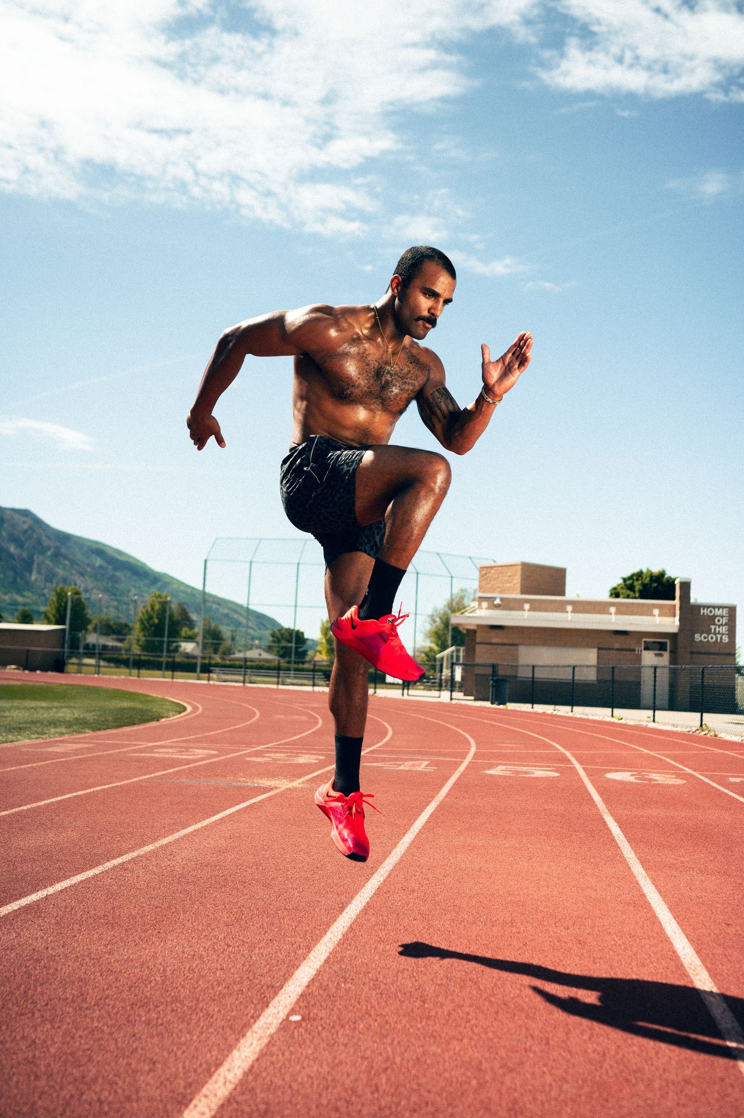 Shirtless male athlete sprinting on a red outdoor track with mountains in the background on a sunny day.