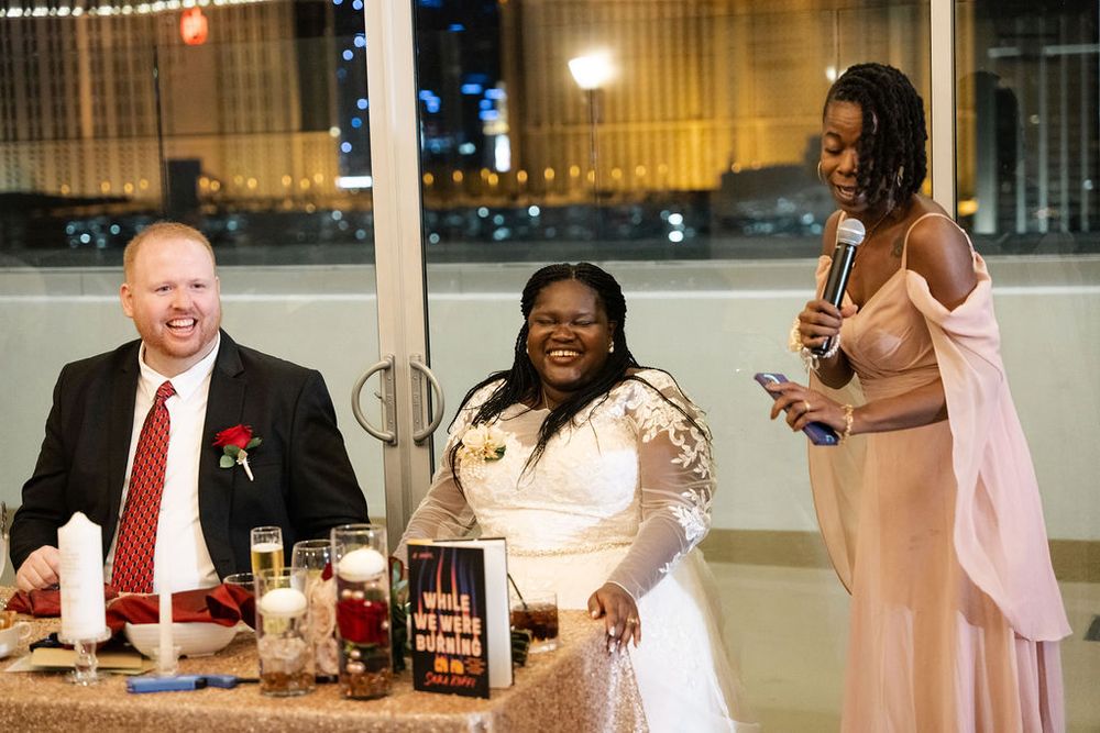 A bride and groom laugh with a bridesmaid giving a speech at an elegant wedding reception dinner.