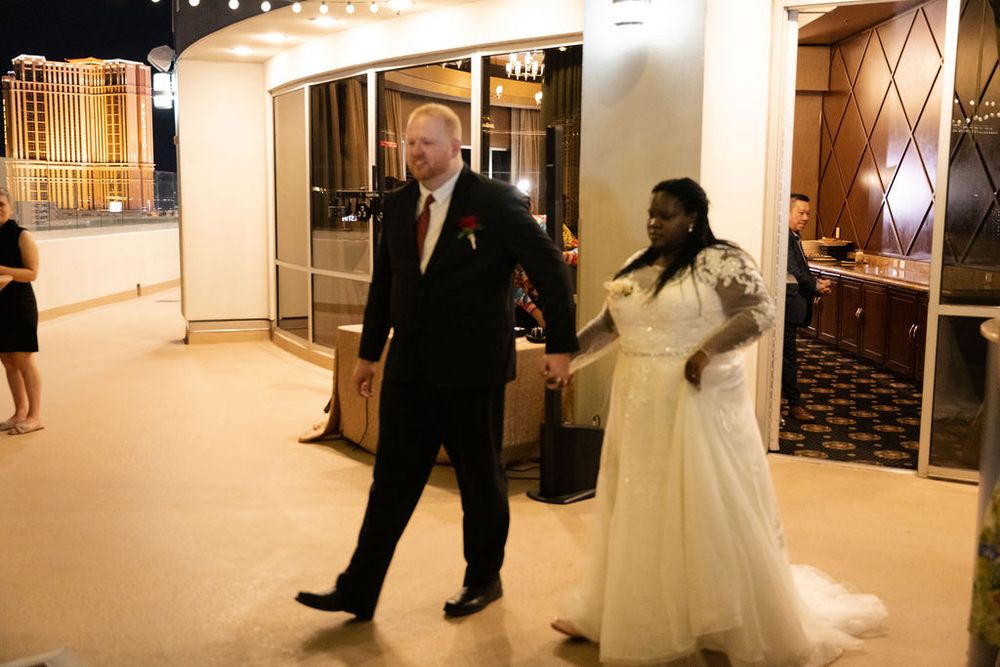 Bride in white gown and groom in black suit hold hands entering wedding reception venue.
