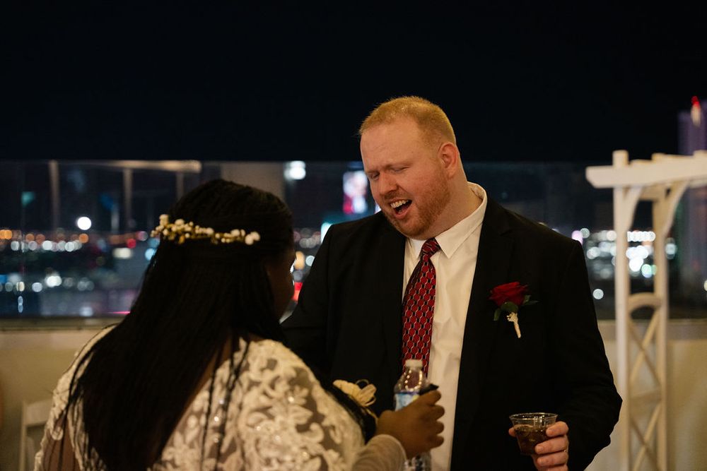 Man in suit with red rose laughs with woman in floral crown at elegant rooftop evening event.