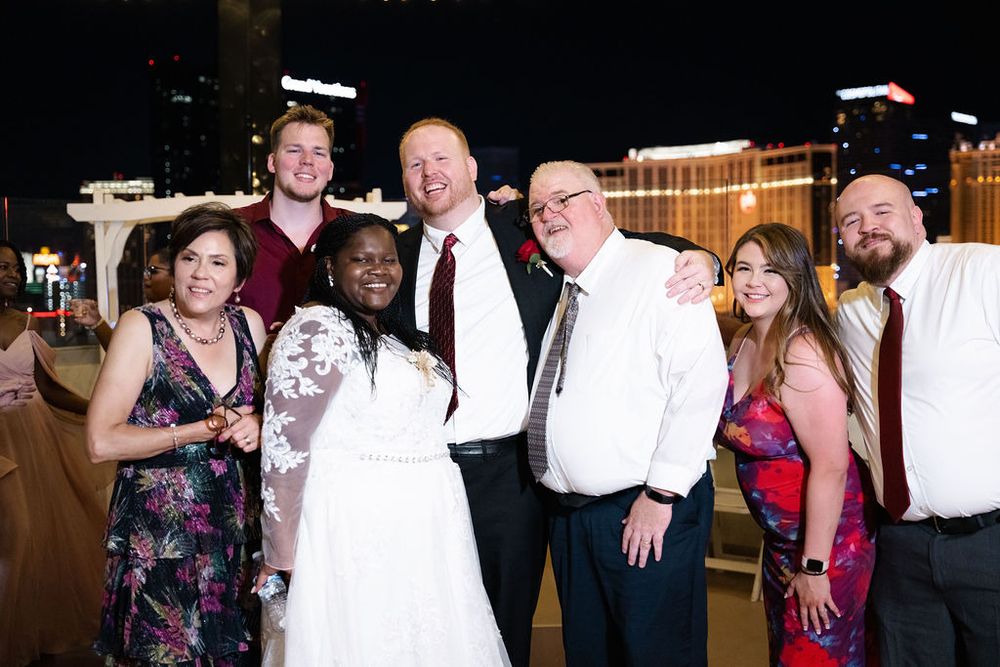 Group of seven people posing together at a Las Vegas wedding celebration at night, with the Strip visible in the background.