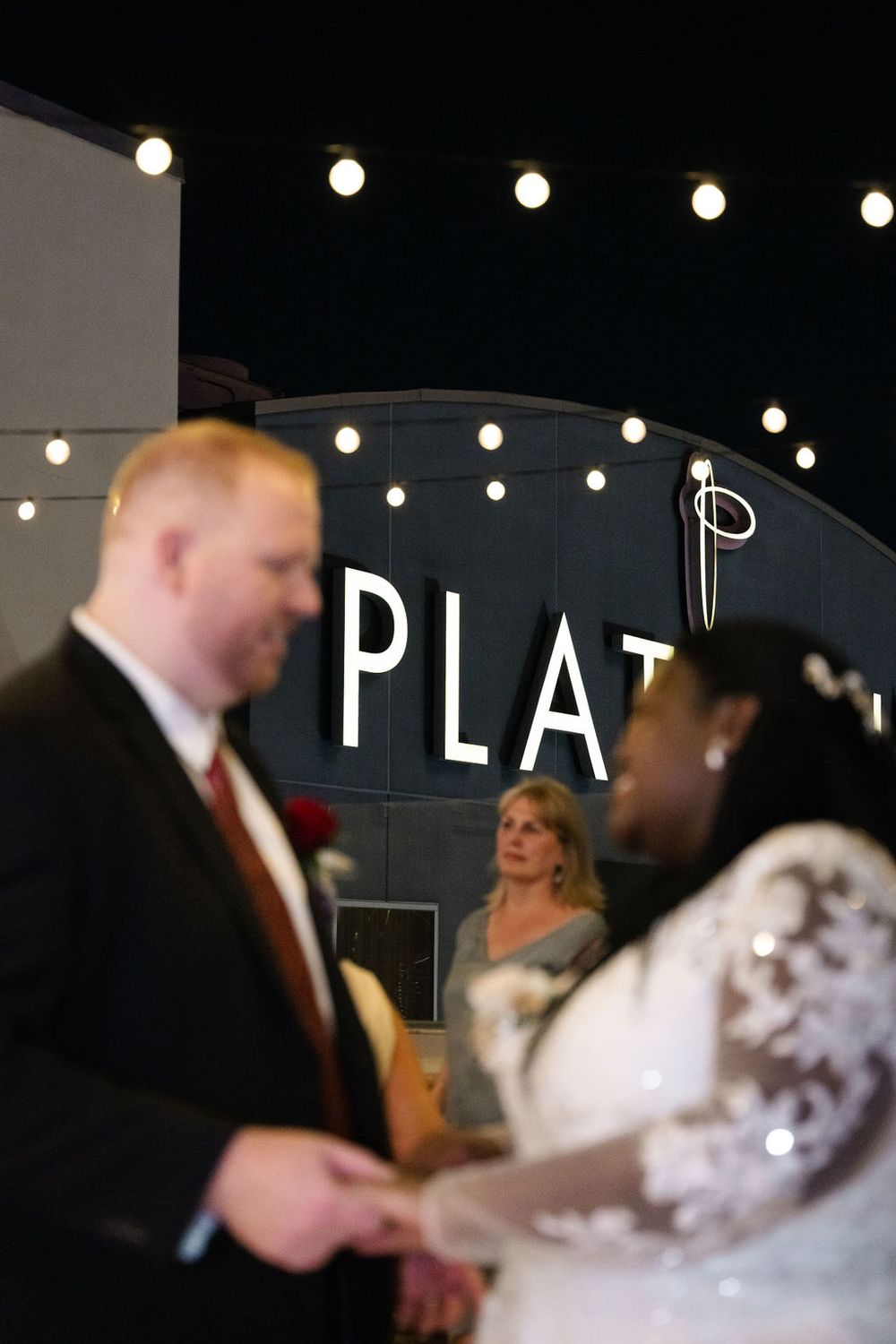 Interracial couple exchanges vows at outdoor wedding ceremony with string lights and PLAT sign backdrop at night.