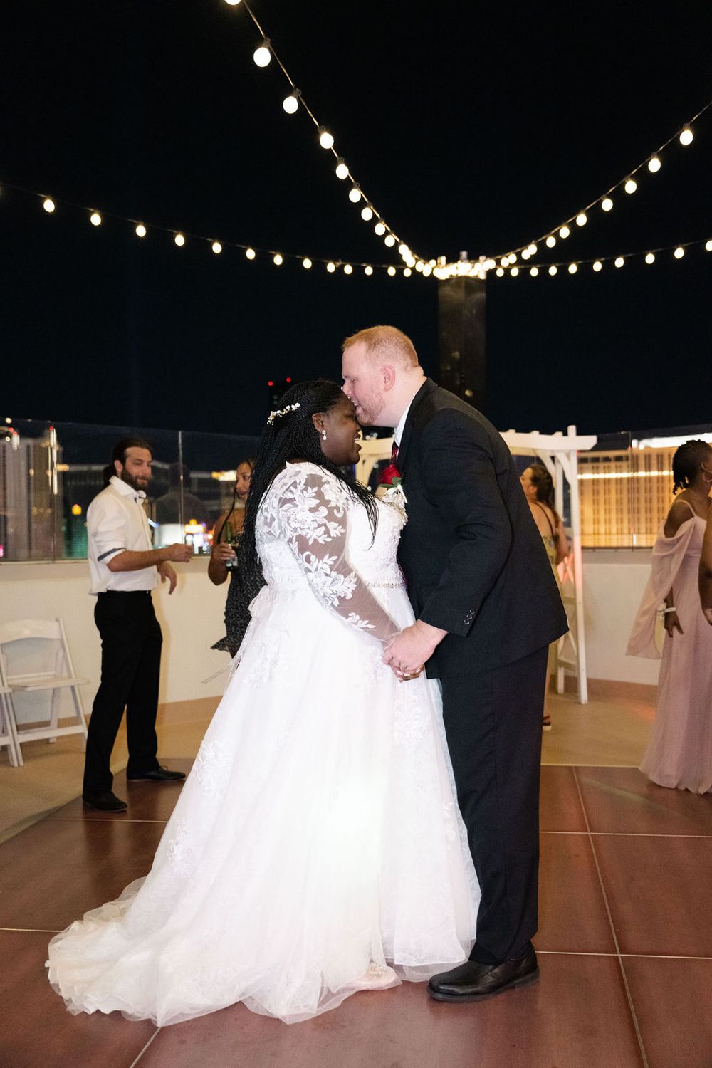 Bride and groom share first dance on outdoor rooftop wedding reception under string lights at night.
