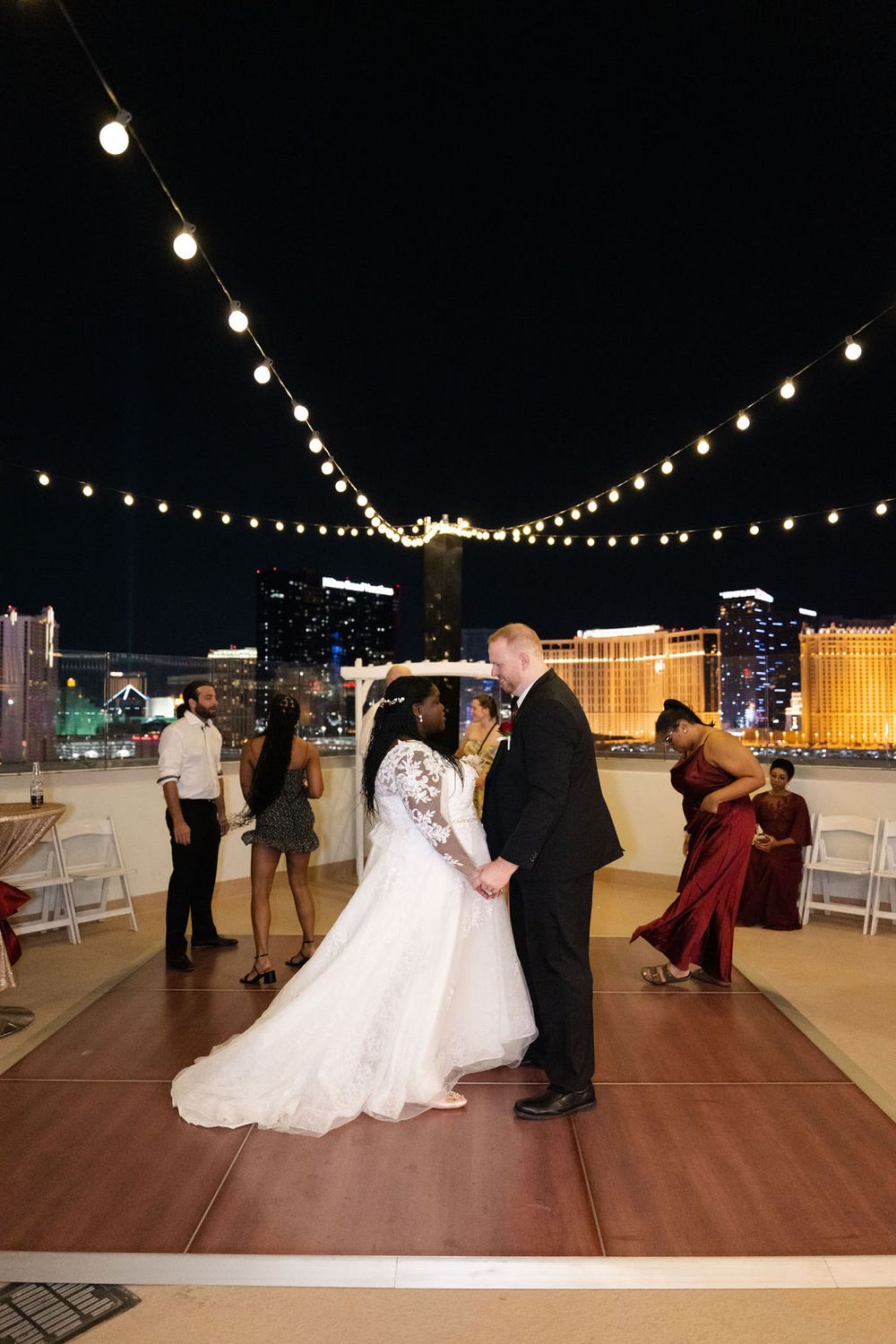 Bride and groom share first dance on rooftop venue with Las Vegas Strip skyline and string lights at night.