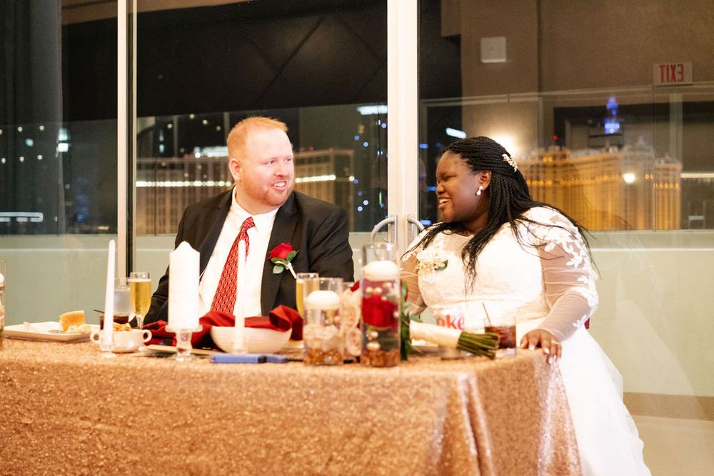 Bride and groom share a joyful moment at their wedding reception sweetheart table with elegant red and gold decor.