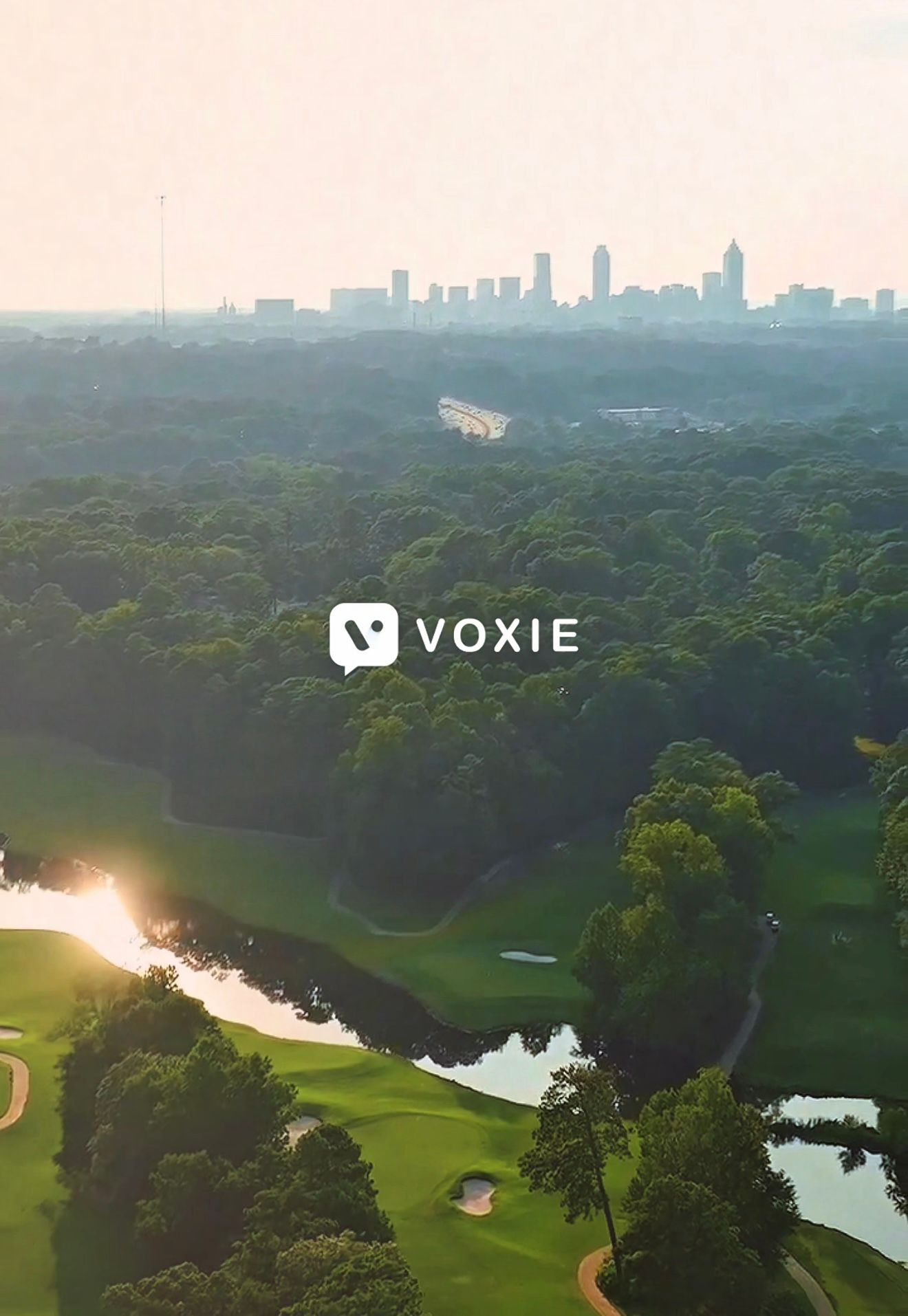Aerial view of lush green golf course with city skyline and Voxie logo overlay at golden hour.
