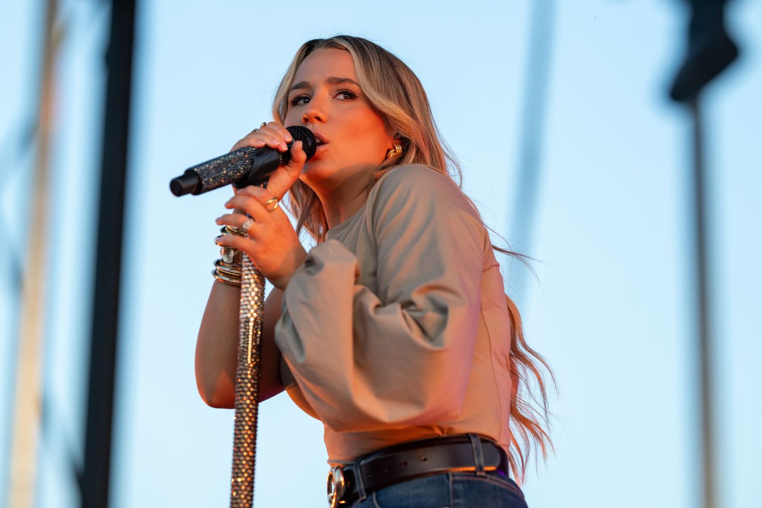 Young female singer performing on outdoor stage, holding microphone, wearing beige top and jeans with long blonde hair.