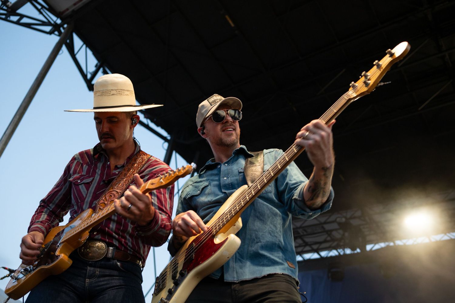 Two male guitarists performing on outdoor stage, one in cowboy hat and flannel, other in denim shirt and cap.