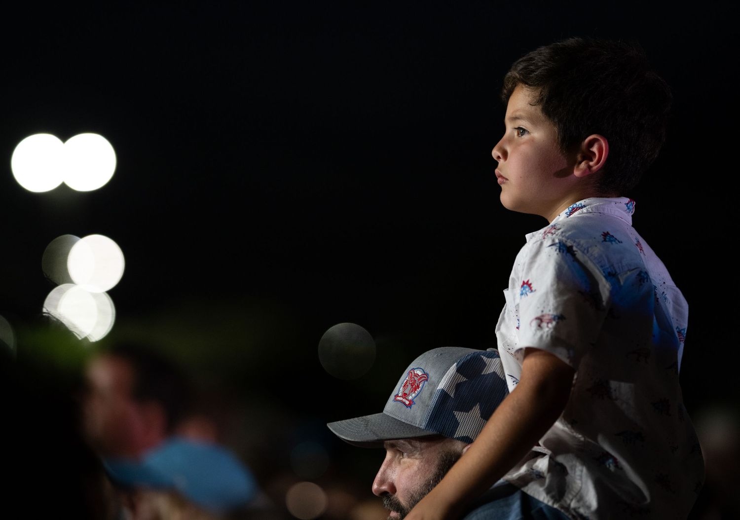 Young boy sitting on shoulders at a nighttime outdoor event, wearing a patterned shirt and white cap.
