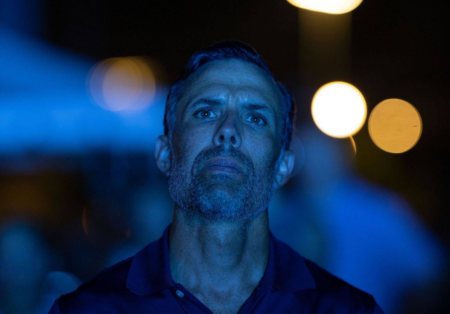 Man with stubble gazing upward in blue-tinted night light with bokeh background.
