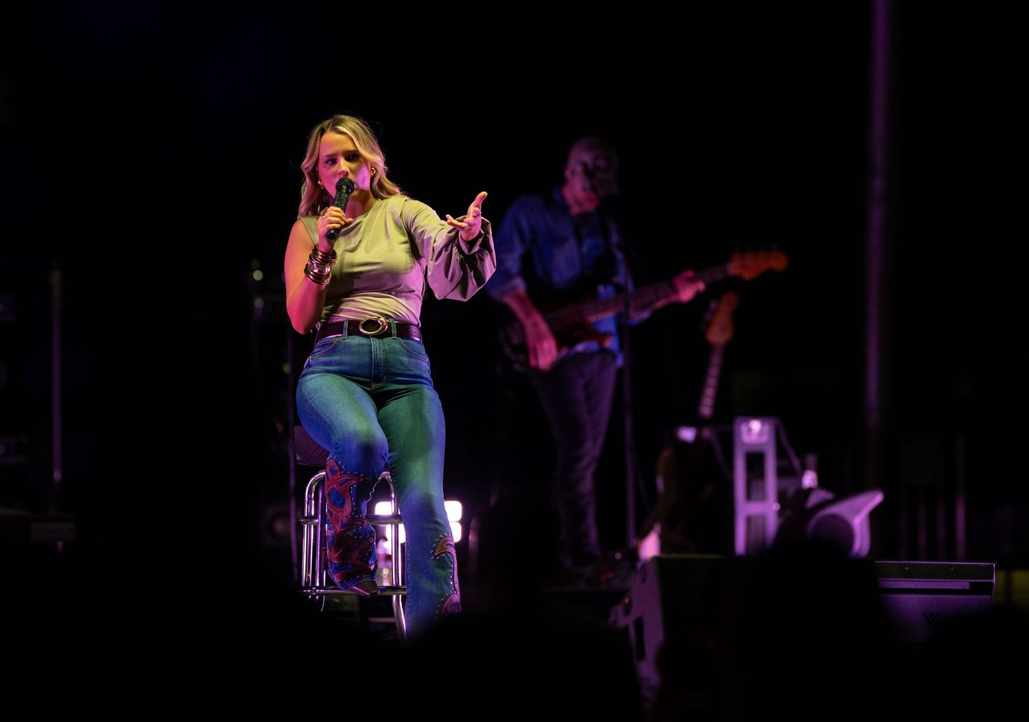 Female singer performing on stage with microphone, wearing green outfit, guitarist in background under colorful lights.