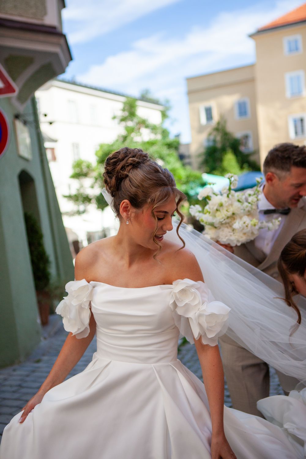 Smiling bride in off-shoulder white gown with floral sleeves stands outdoors on cobblestone street on her wedding day.