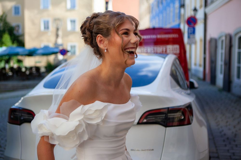 Laughing bride in white off-shoulder gown and veil stands beside a white car on a European cobblestone street.