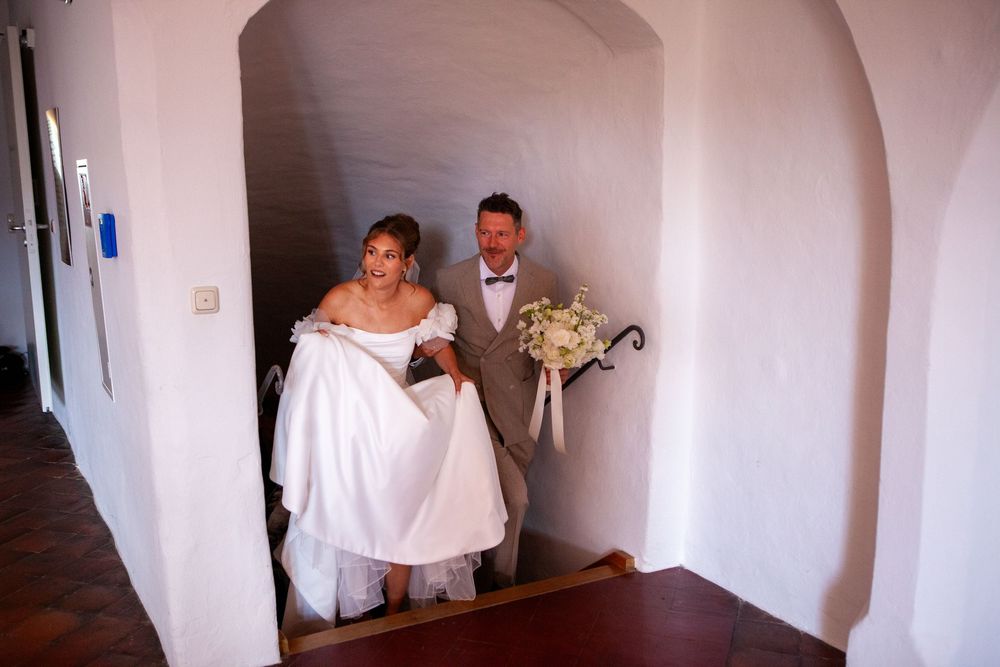 A bride in a white gown and groom holding a bouquet walk up stairs in a whitewashed venue on their wedding day.