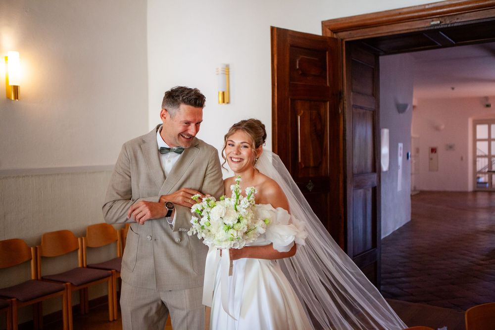 Happy bride and groom sharing a joyful moment indoors, bride holding white bouquet in elegant wedding attire.