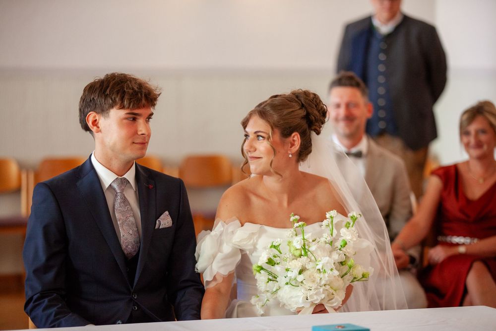 Bride and groom sitting together at wedding ceremony, bride holding white bouquet, guests seated in background.